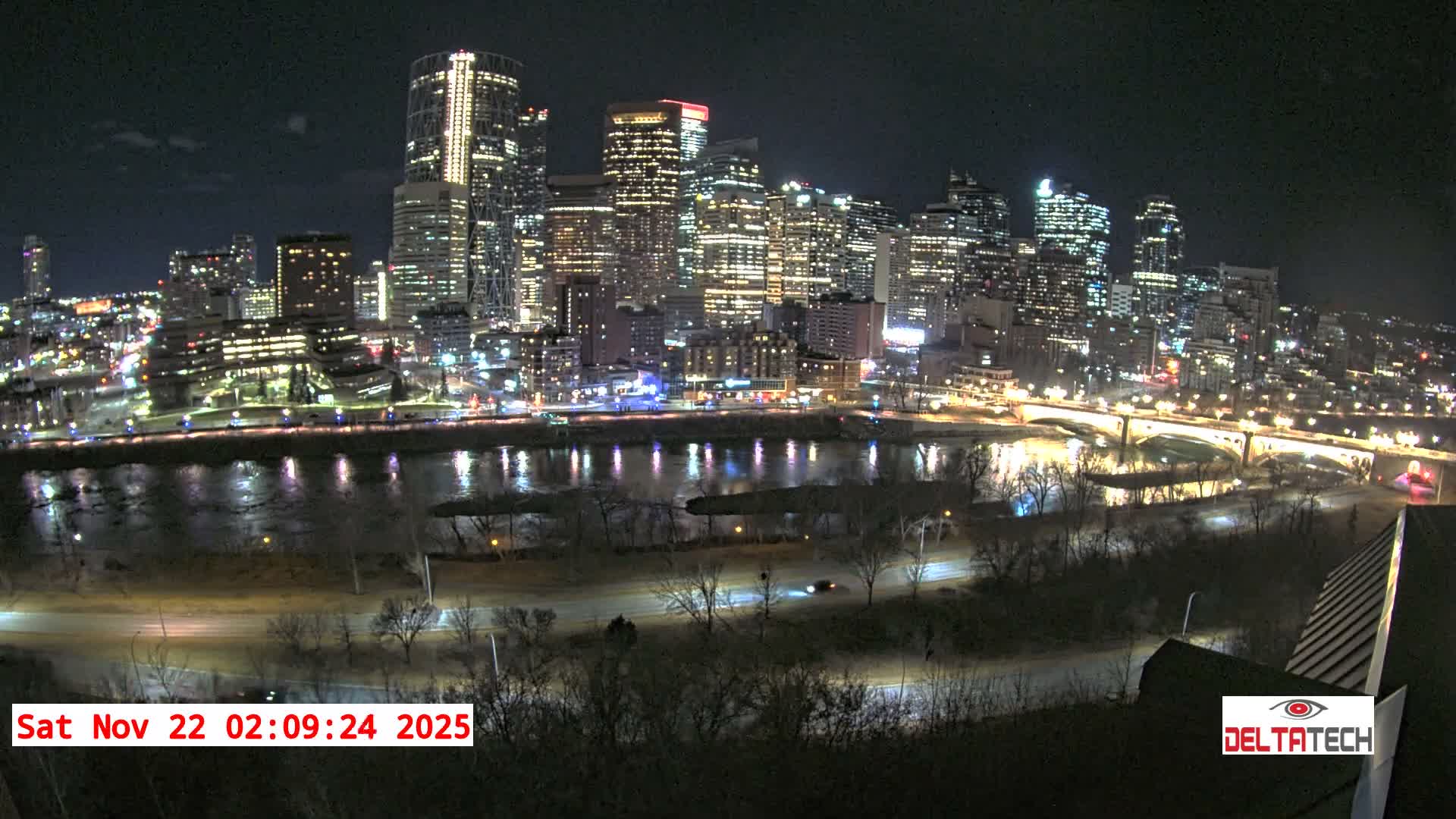 A nighttime cityscape features numerous illuminated buildings reflecting in a wide river, with bridges, roads, and bare trees along the banks under clear skies.