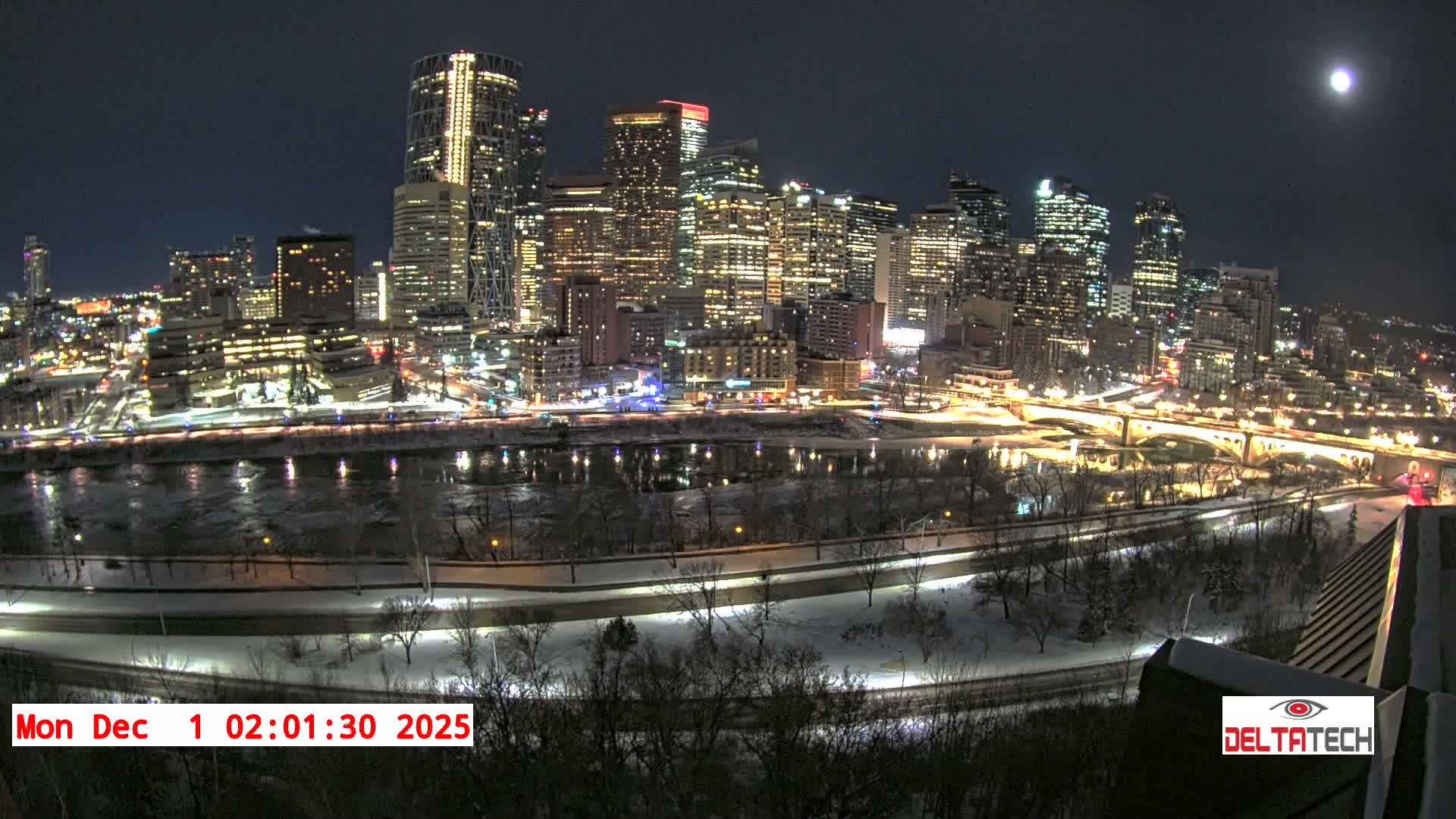 A vibrant city skyline illuminates a clear, cold winter night, reflecting across a partially frozen river bordered by snow-covered banks and roads.