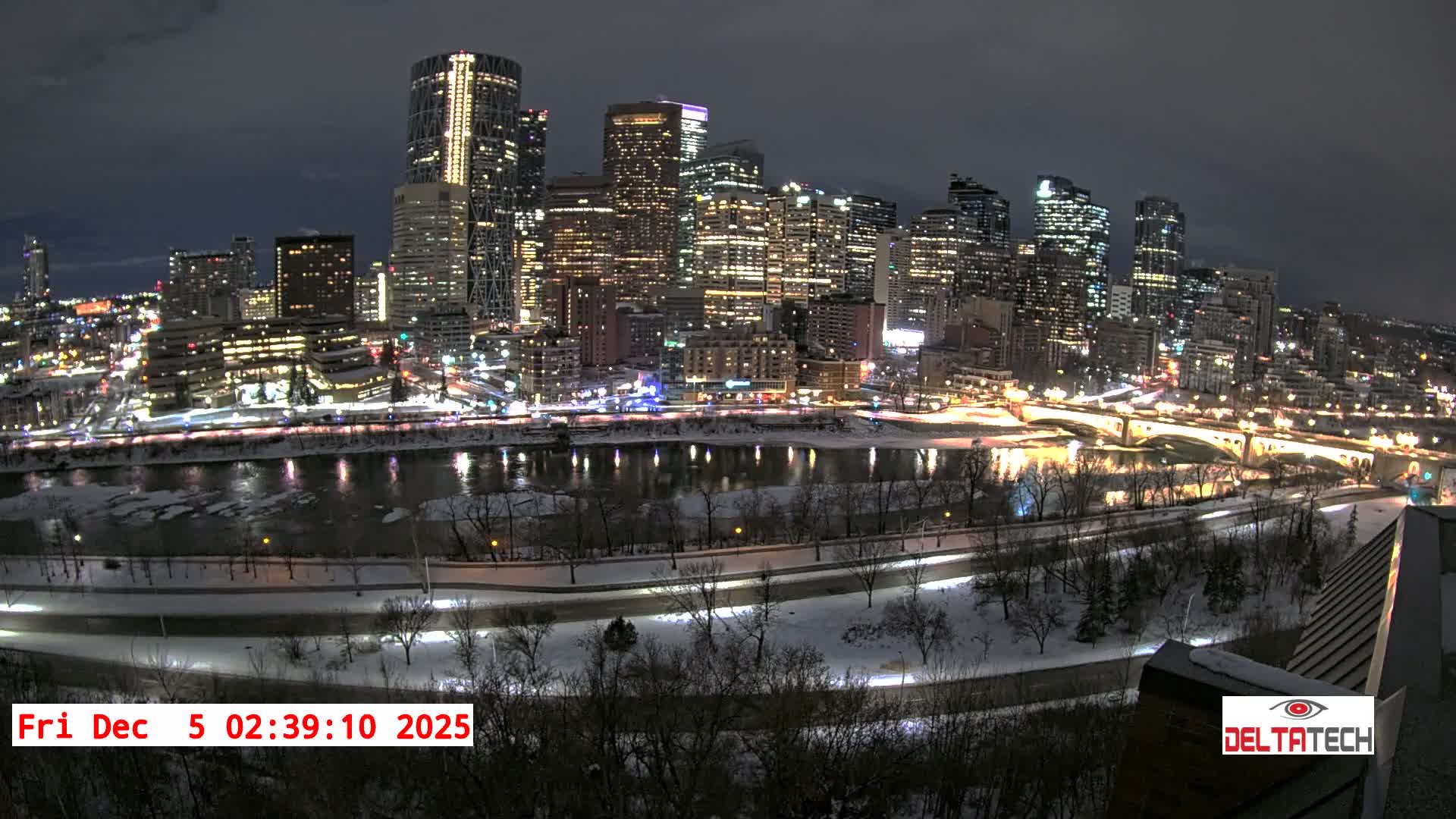 A panoramic nighttime cityscape reveals a brightly lit skyline of skyscrapers reflected in a partially frozen river, flanked by snow-covered banks and roads, under a dark, cloudy winter sky.