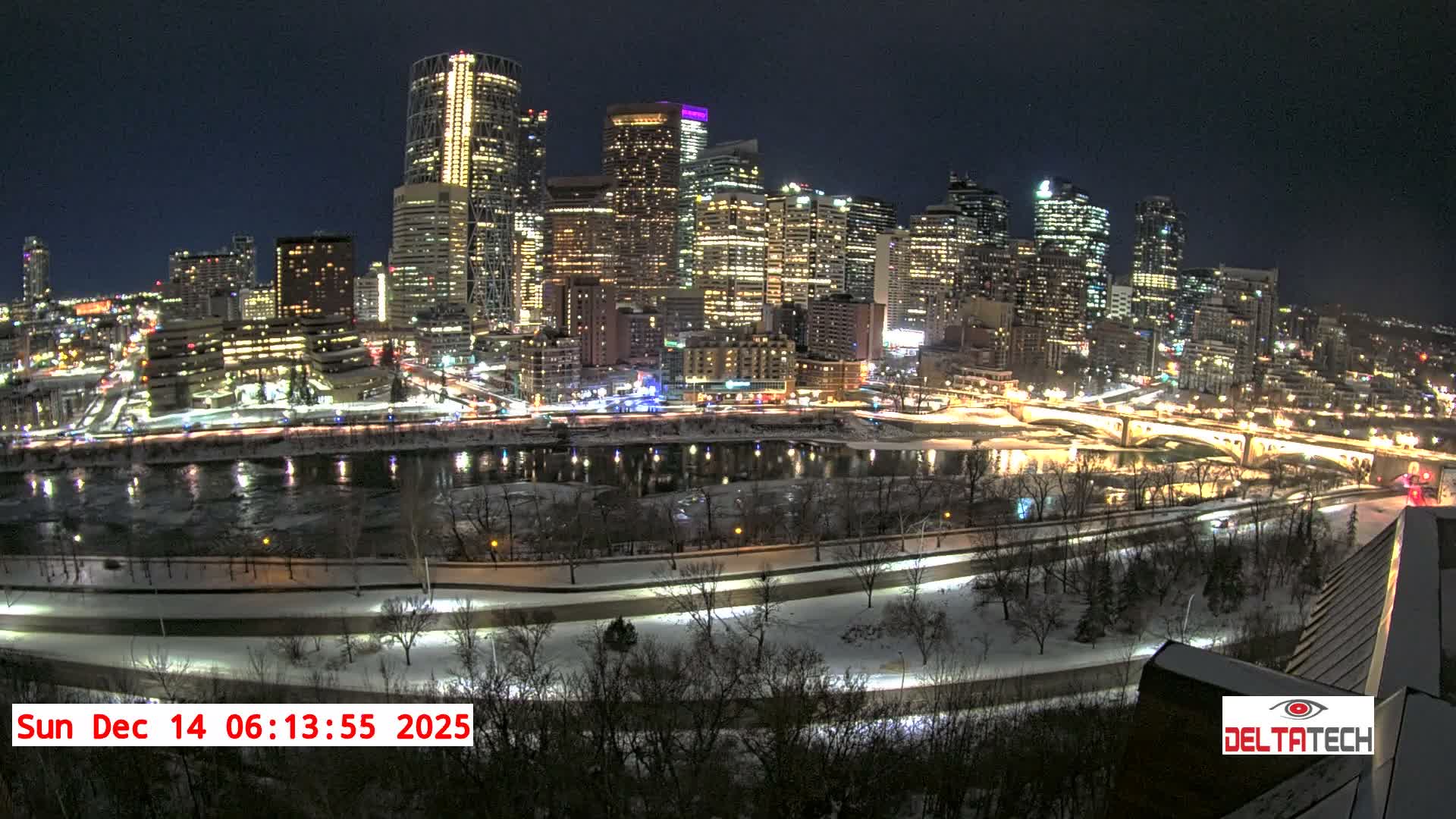 A panoramic nighttime cityscape reveals a brightly lit skyline of skyscrapers reflected in a partially frozen river, flanked by snow-covered banks and roads, under a dark, cloudy winter sky.