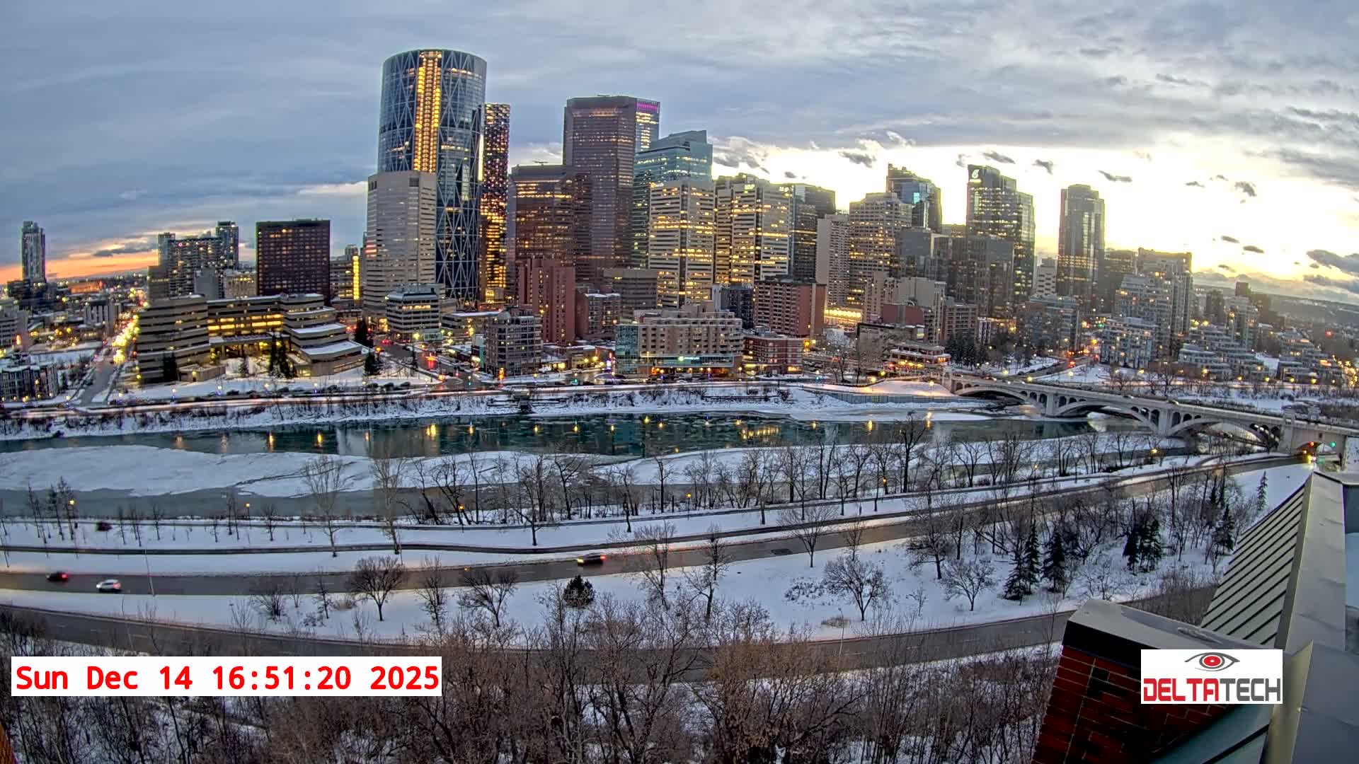 A panoramic nighttime cityscape reveals a brightly lit skyline of skyscrapers reflected in a partially frozen river, flanked by snow-covered banks and roads, under a dark, cloudy winter sky.