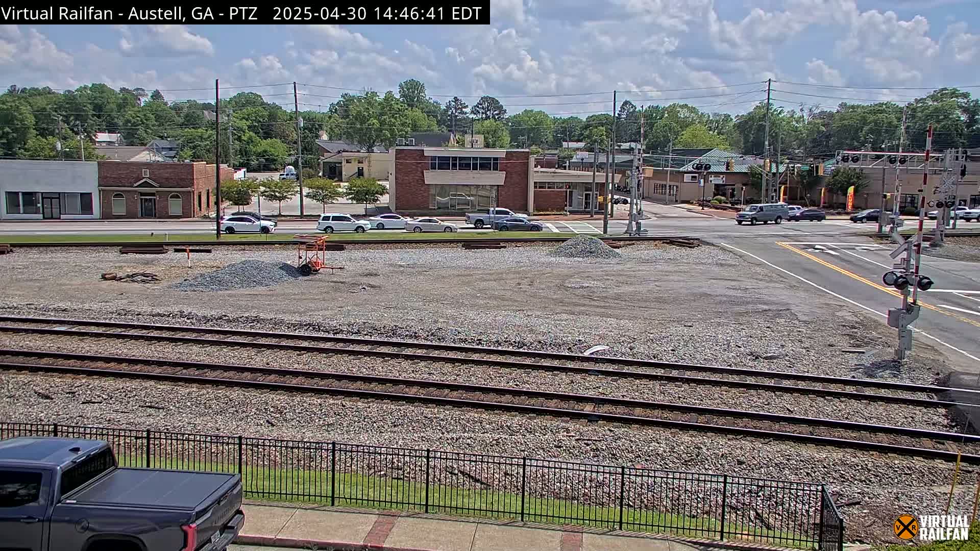 A partly sunny day reveals a railroad crossing near a town's street intersection, with several cars at the crossing and piles of gravel in the foreground.