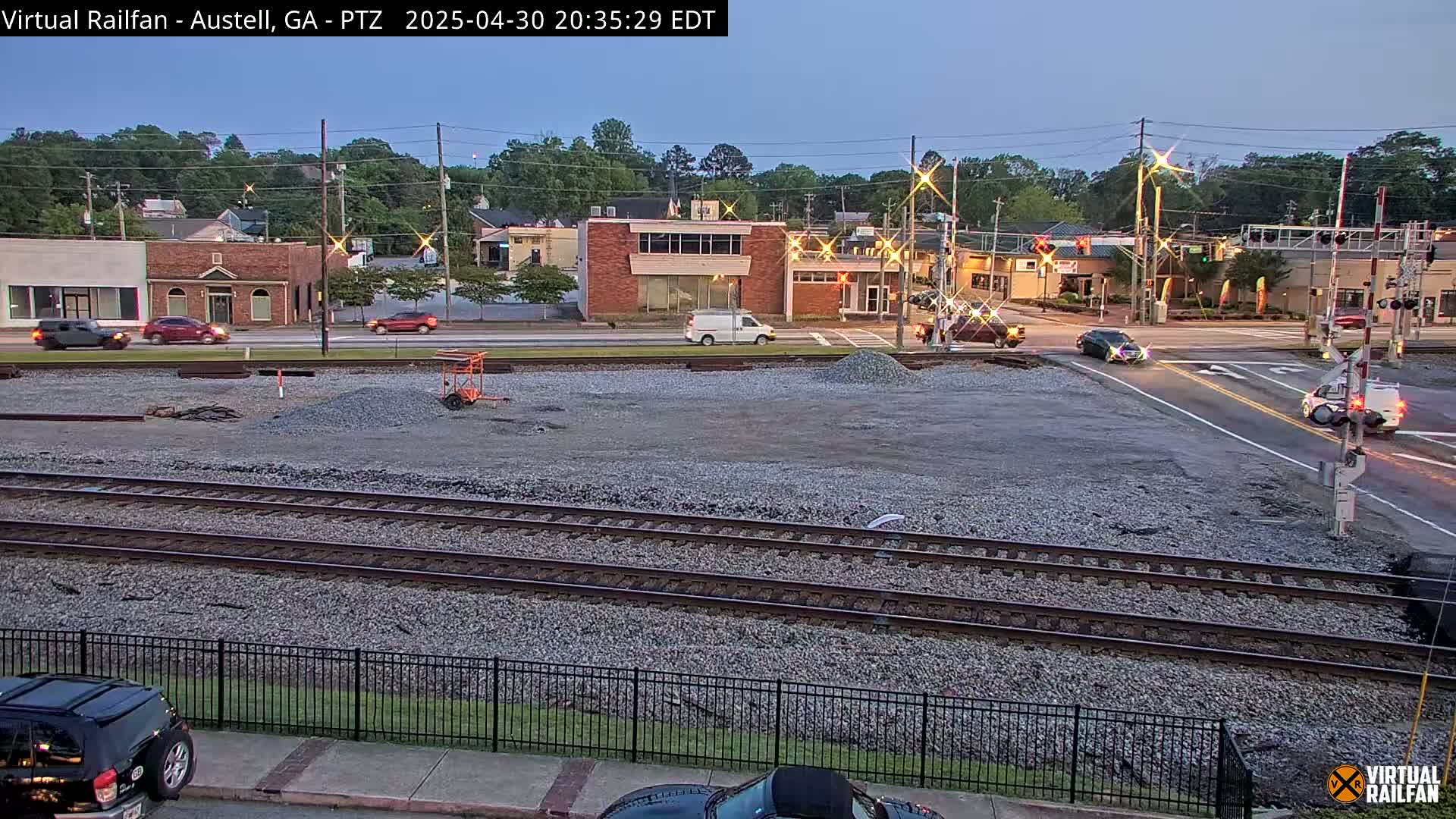 A twilight view of a street intersection with railroad tracks in the foreground, showing several vehicles and buildings in the background under a clear sky.