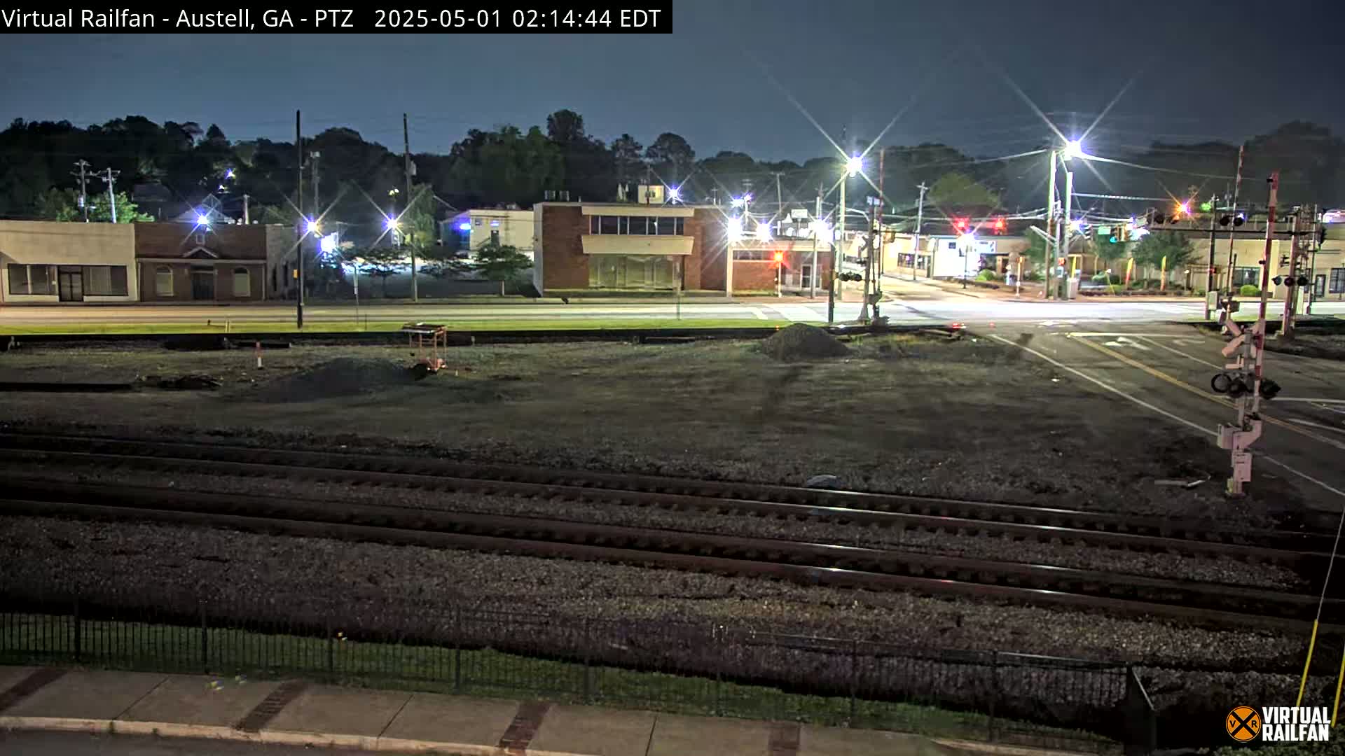 At night, a mostly empty street intersects with railroad tracks, bordered by low-rise buildings and streetlights under a clear sky.