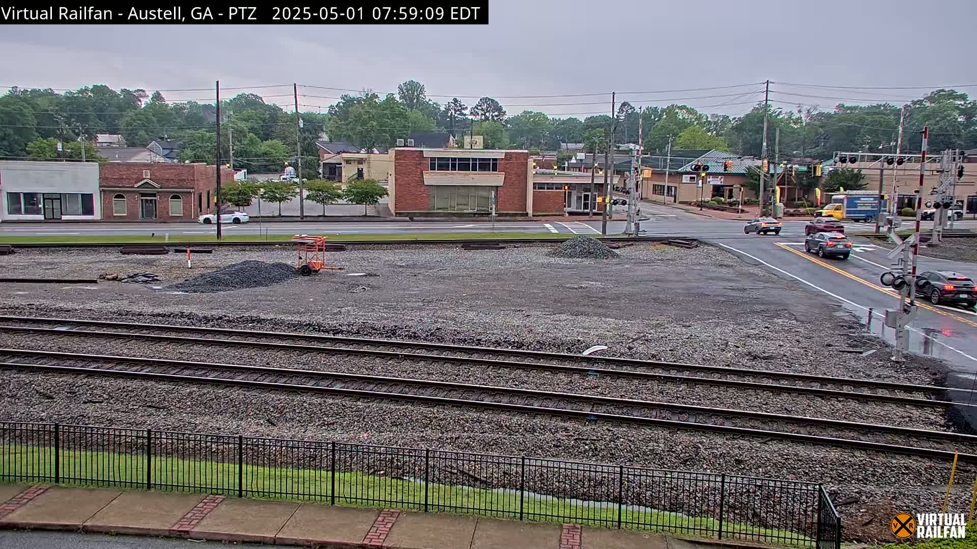 A view of a gravel lot next to train tracks, with several buildings and cars visible in the background under an overcast sky.