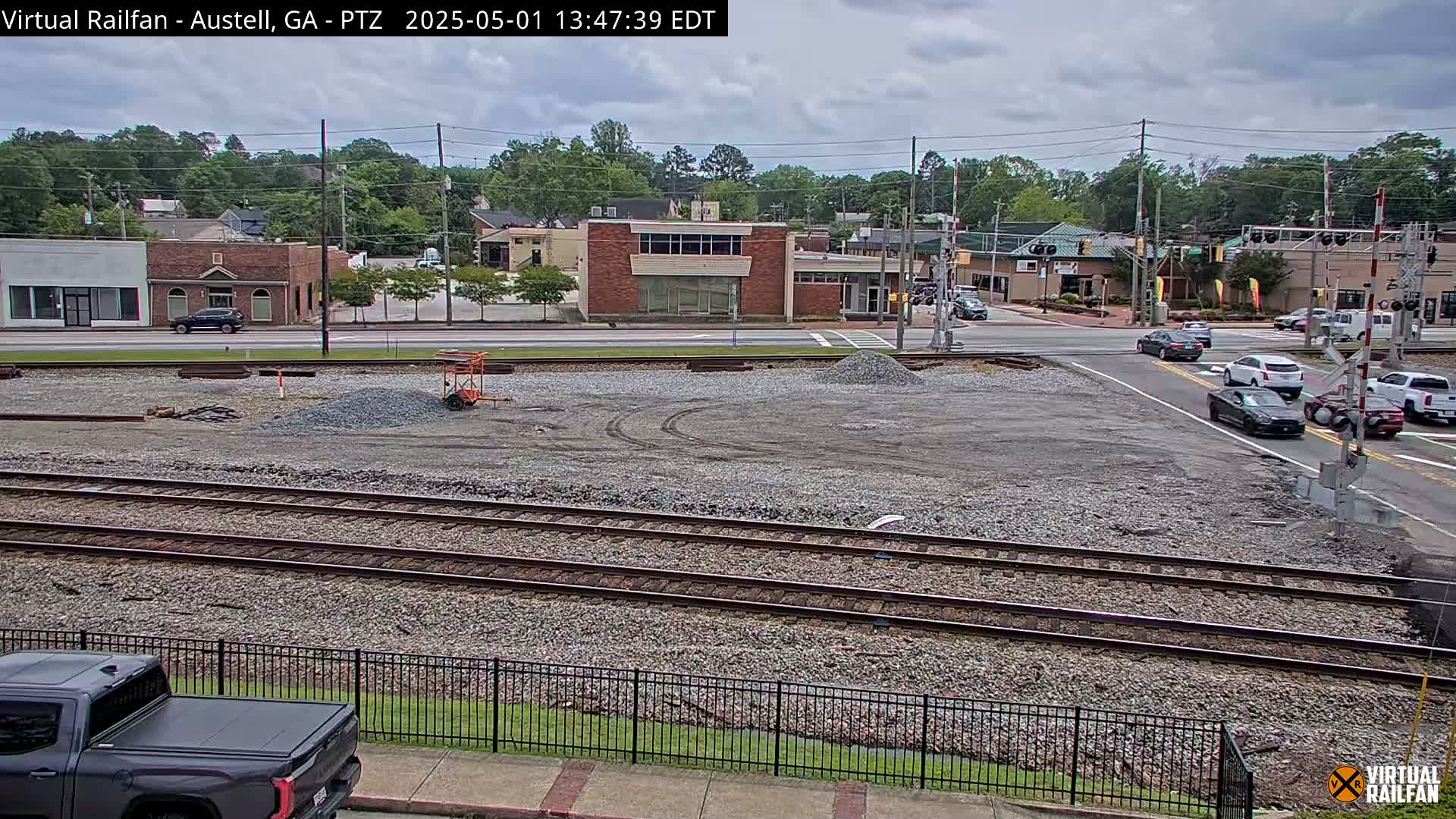 A mostly-cloudy day shows a view of railroad tracks next to a road intersection with several cars, a construction area, and buildings in the background.