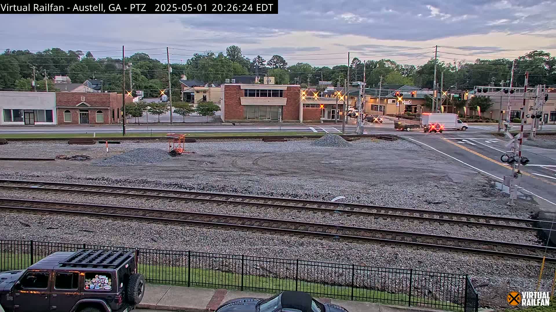 A view of railroad tracks next to a road intersection in front of low-rise buildings under a twilight sky.