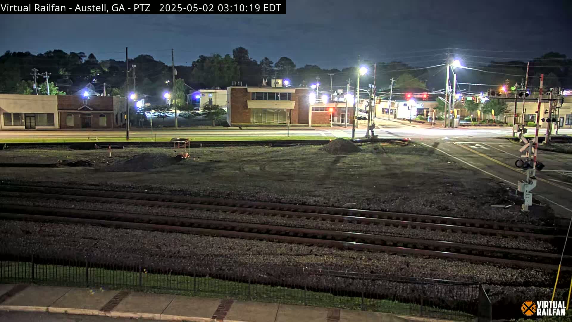 A nighttime view of a railroad crossing near a street with several buildings, under a clear, dark sky.