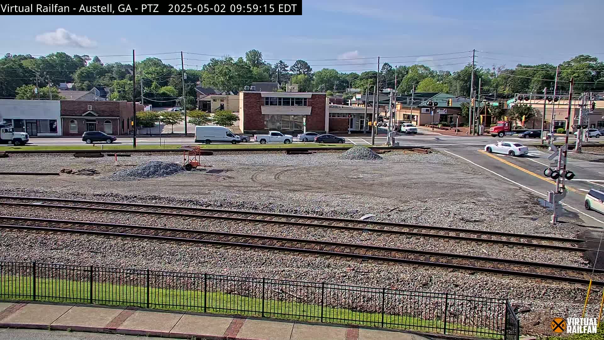 A sunny day reveals a railway crossing near a town's commercial area, with several cars visible on the road and piles of gravel in a construction or vacant lot.