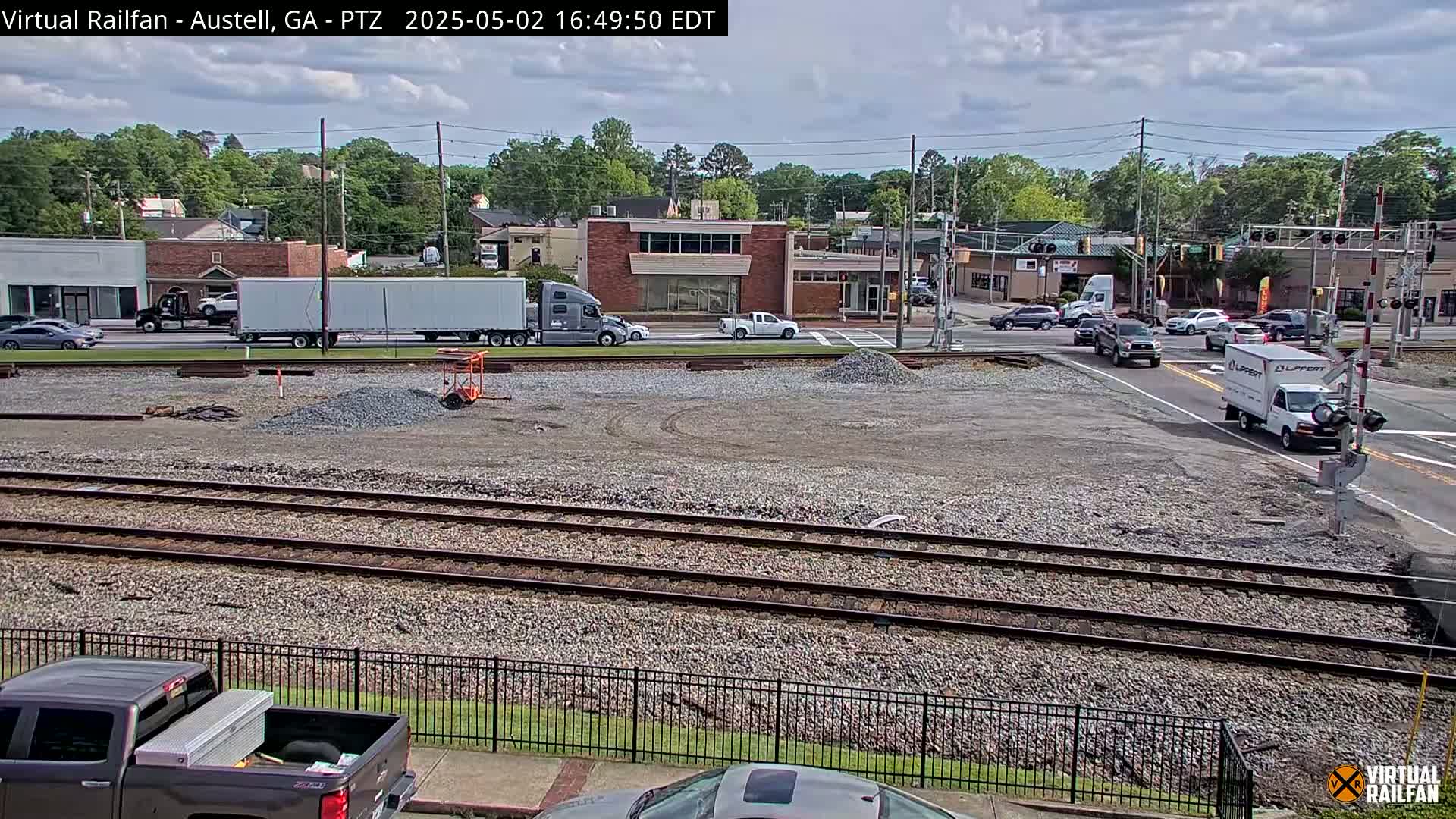 Under a partly cloudy sky, a mostly empty construction site with railroad tracks is shown, next to a road with several vehicles, including semi-trucks and smaller delivery vans.