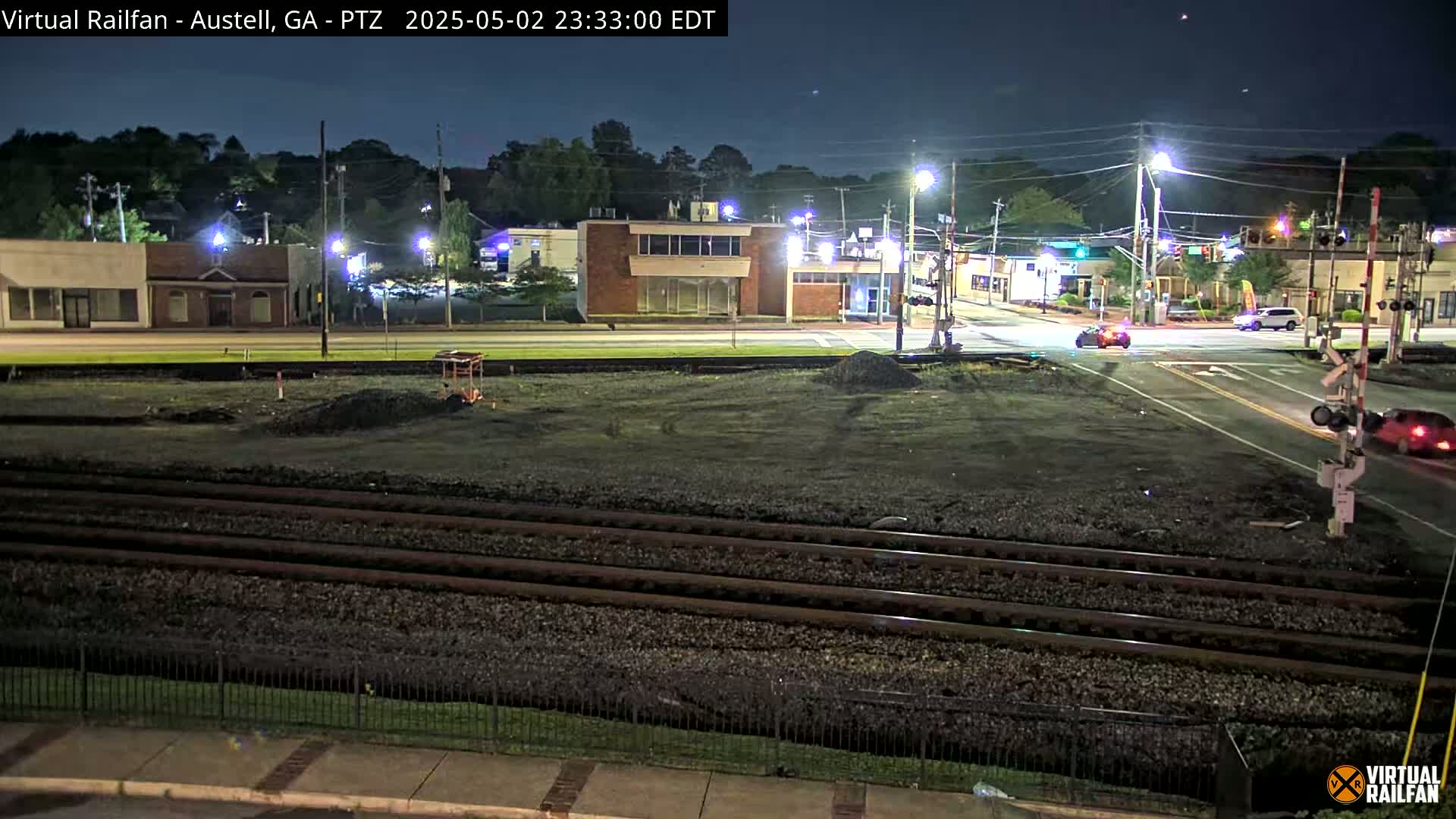 A nighttime view of a street intersection near railroad tracks, with several buildings and a few cars visible under a clear, dark sky.