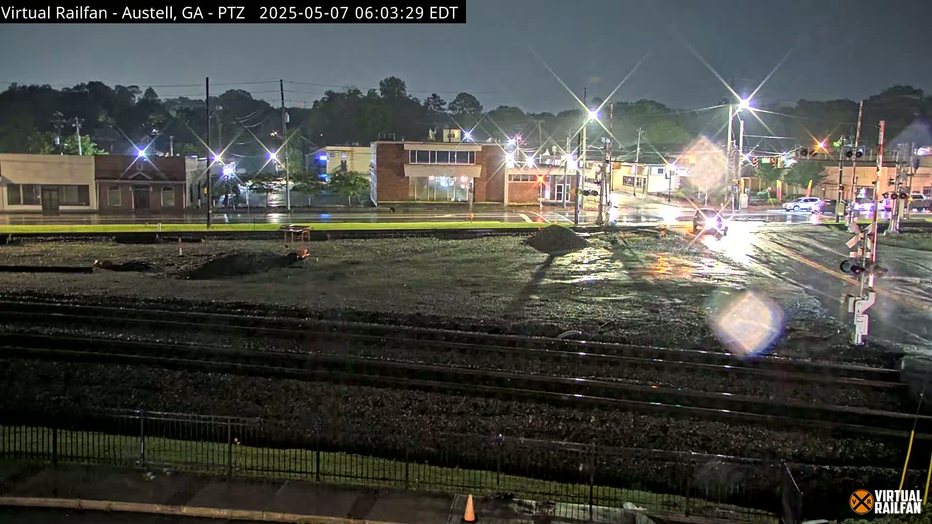 A nighttime view of a rain-slicked street intersection near railroad tracks, with several buildings visible in the background.