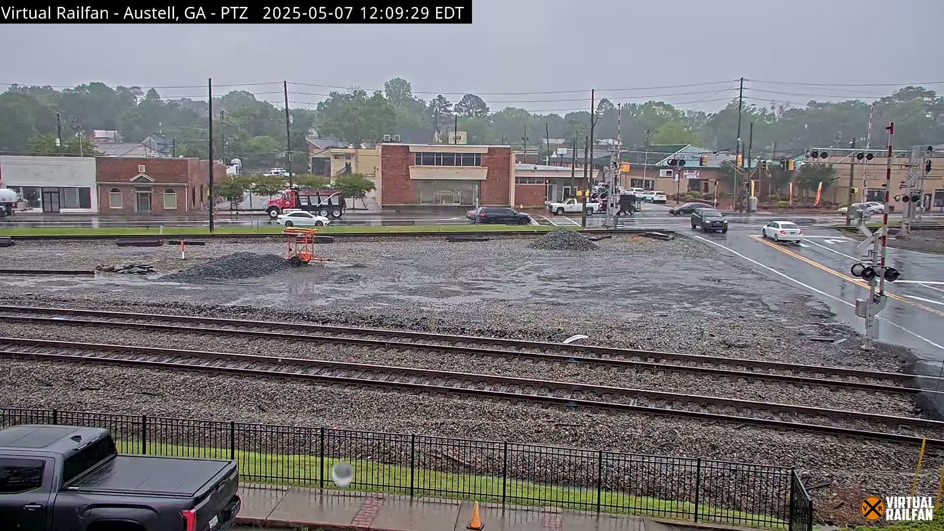 A rain-soaked view of railroad tracks in a town, with several vehicles at a nearby intersection and piles of gravel on the tracks.