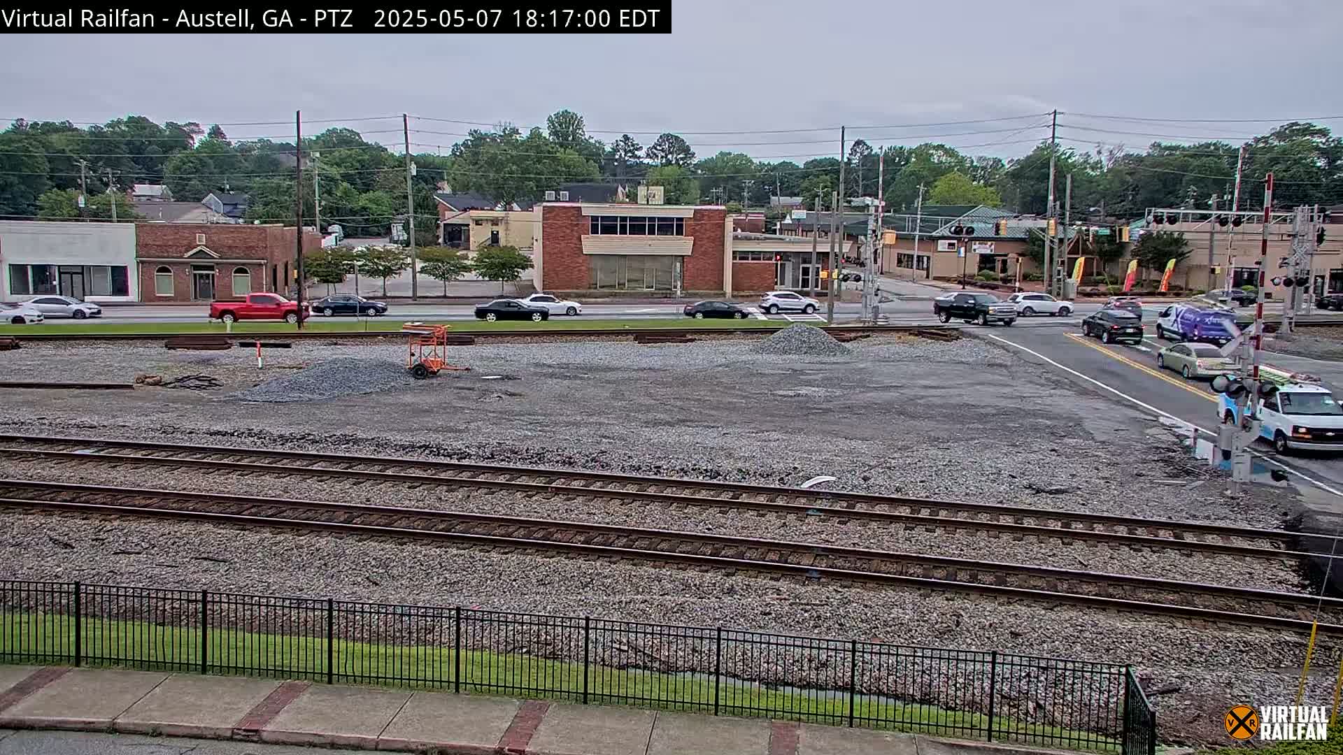 A mostly cloudy day shows a railway crossing in front of a small town with several cars at the intersection and a large pile of gravel in the foreground.