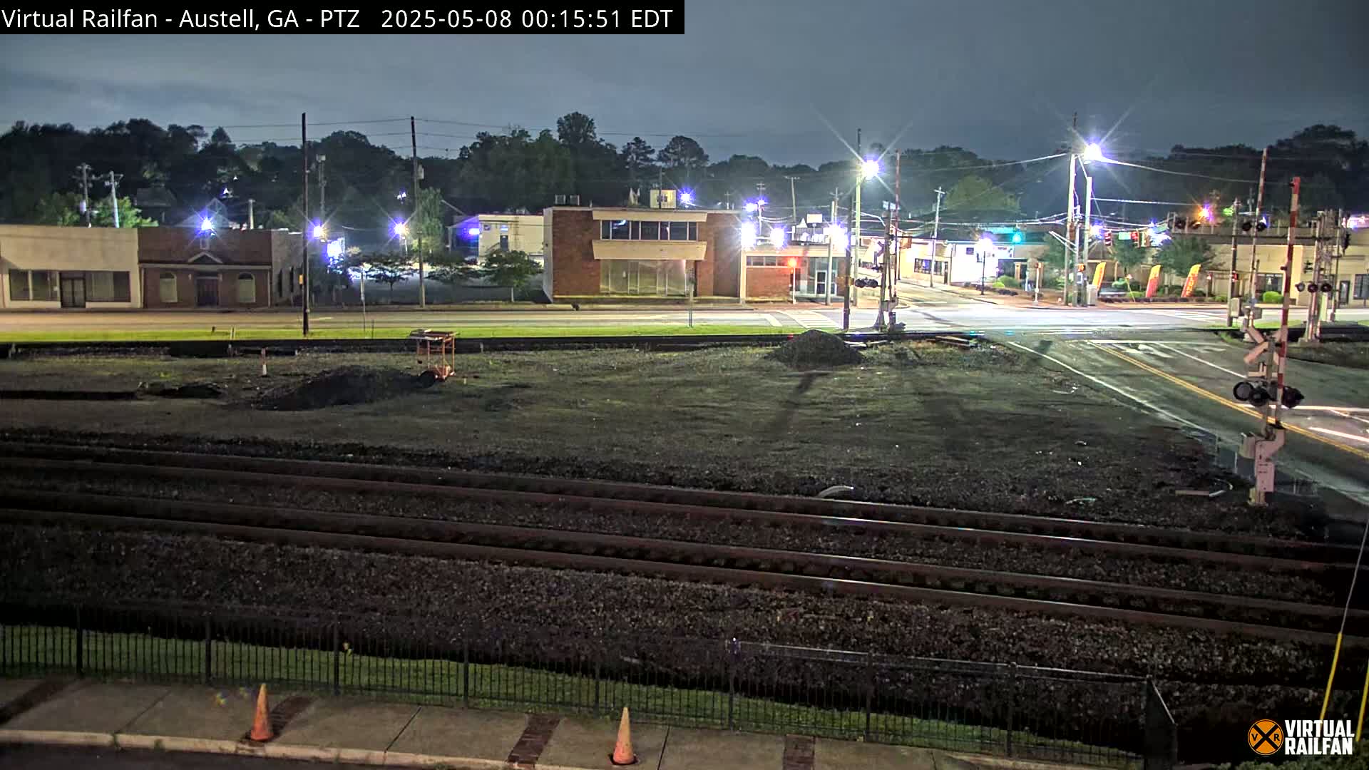 Under a dark night sky, a nighttime view shows a railway crossing near a street intersection with several buildings and streetlights, and construction in progress near the tracks.