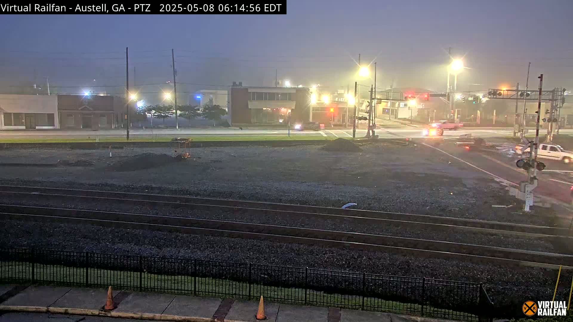 A misty night scene shows railroad tracks in front of a street intersection with cars and buildings in the background.