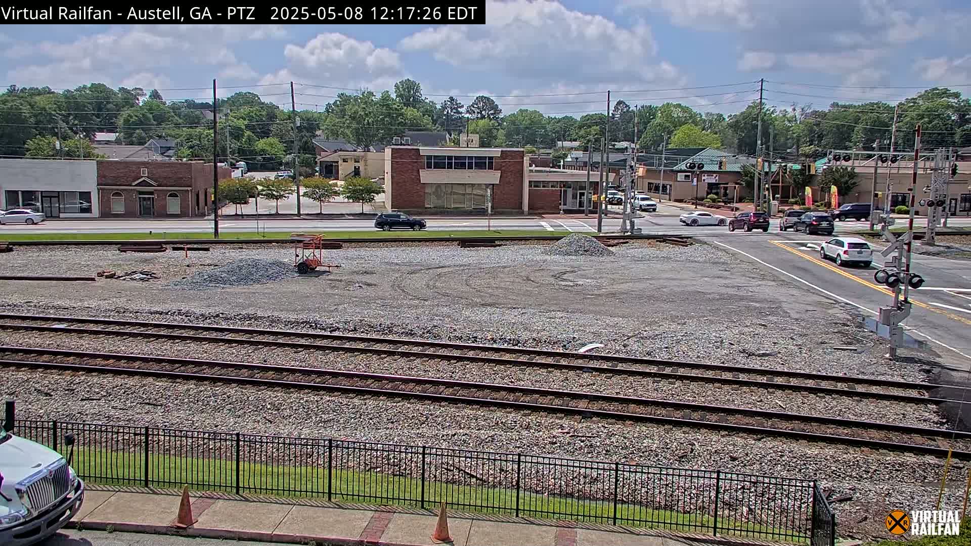 A mostly sunny day reveals a railway crossing near a town's buildings and a construction site, with several cars visible at an intersection.