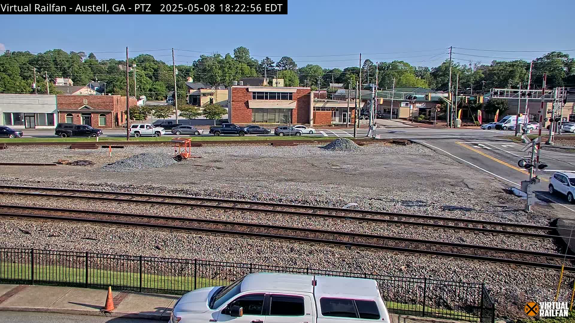 A sunny day view of a railroad crossing in a town with several cars and buildings, and a construction site with gravel in the foreground.