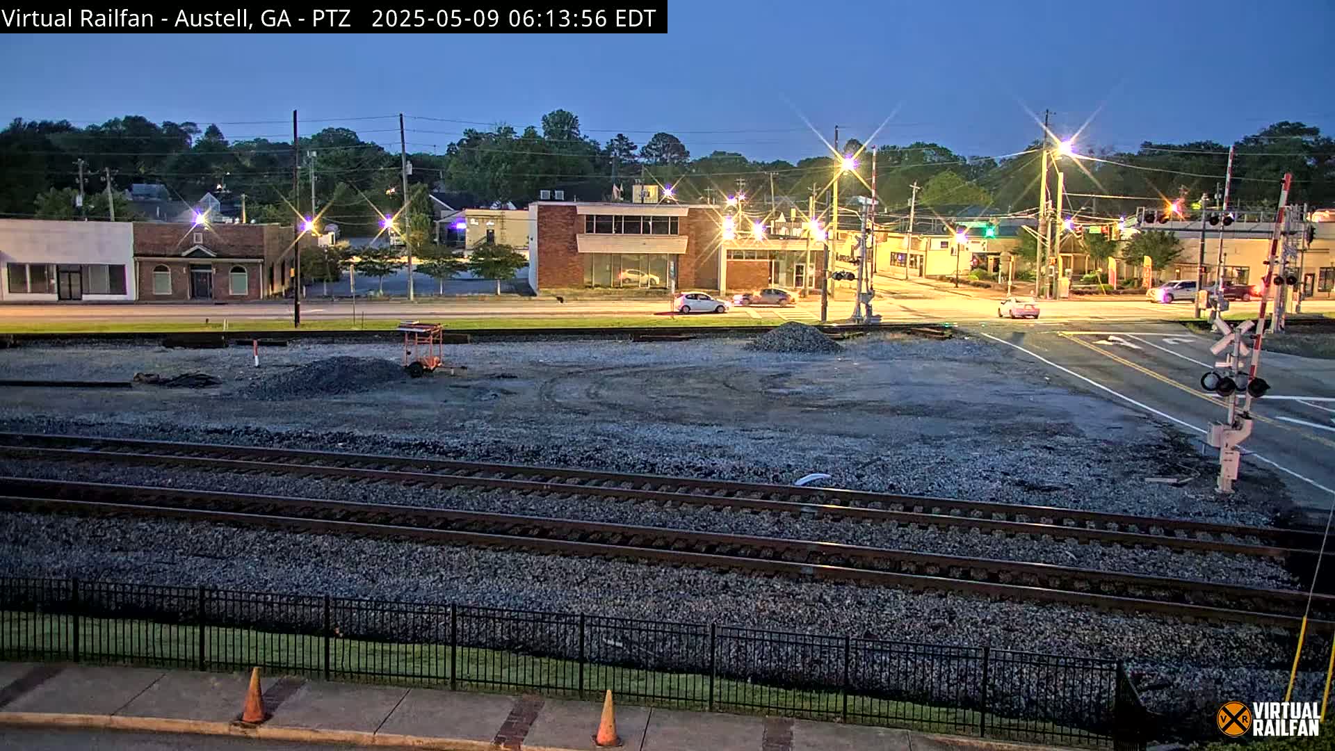 A twilight view of train tracks next to a road and buildings, with a few cars visible and construction material in the foreground.