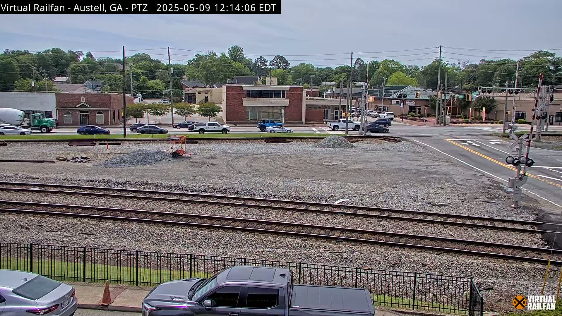 A mostly clear day shows a view of railroad tracks next to a paved road intersection, with several vehicles visible and construction materials on a cleared lot nearby, a few buildings are visible in the background.