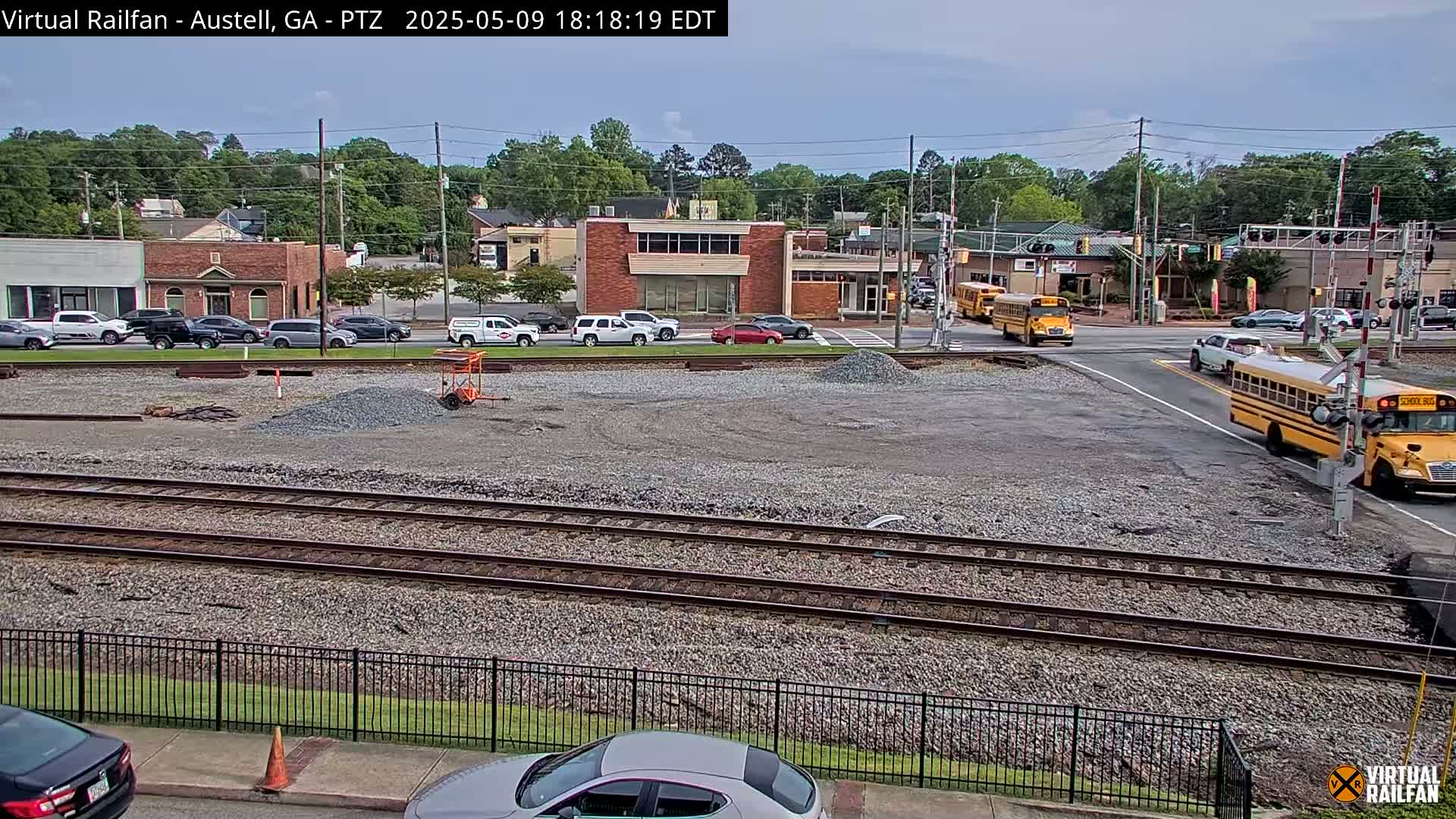 A mostly sunny view of a town's railway crossing, showing several cars and two school buses, with a gravel area in the foreground undergoing construction.