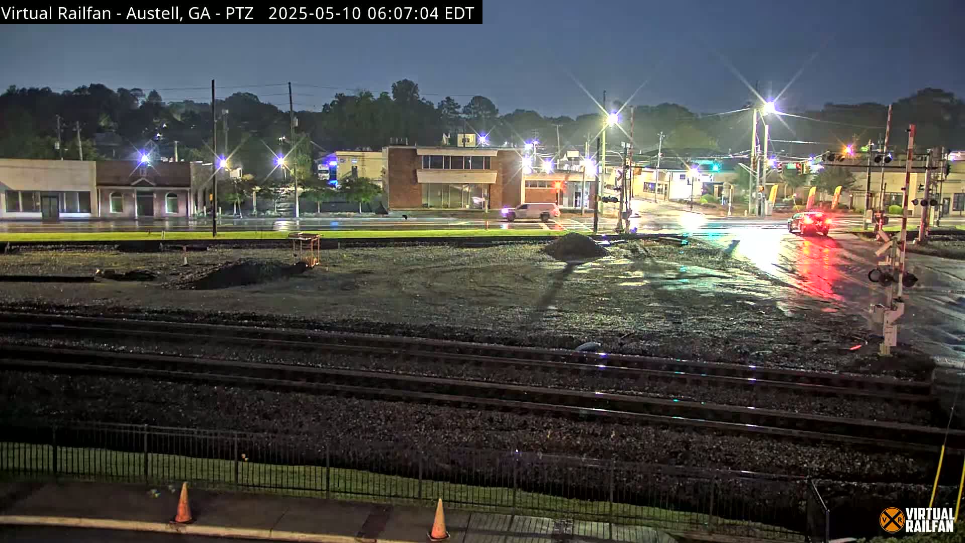 A wet, nighttime street scene shows a railroad crossing near a few buildings under streetlights, with a few vehicles visible on the road.