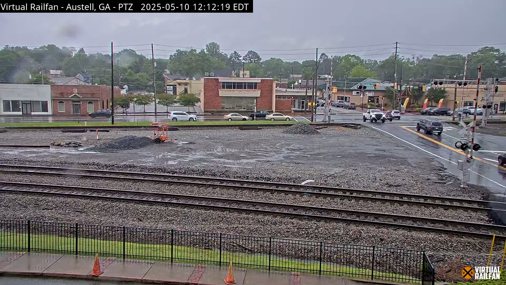 A wet, overcast scene shows railroad tracks and a street intersection with several cars and buildings in the background.