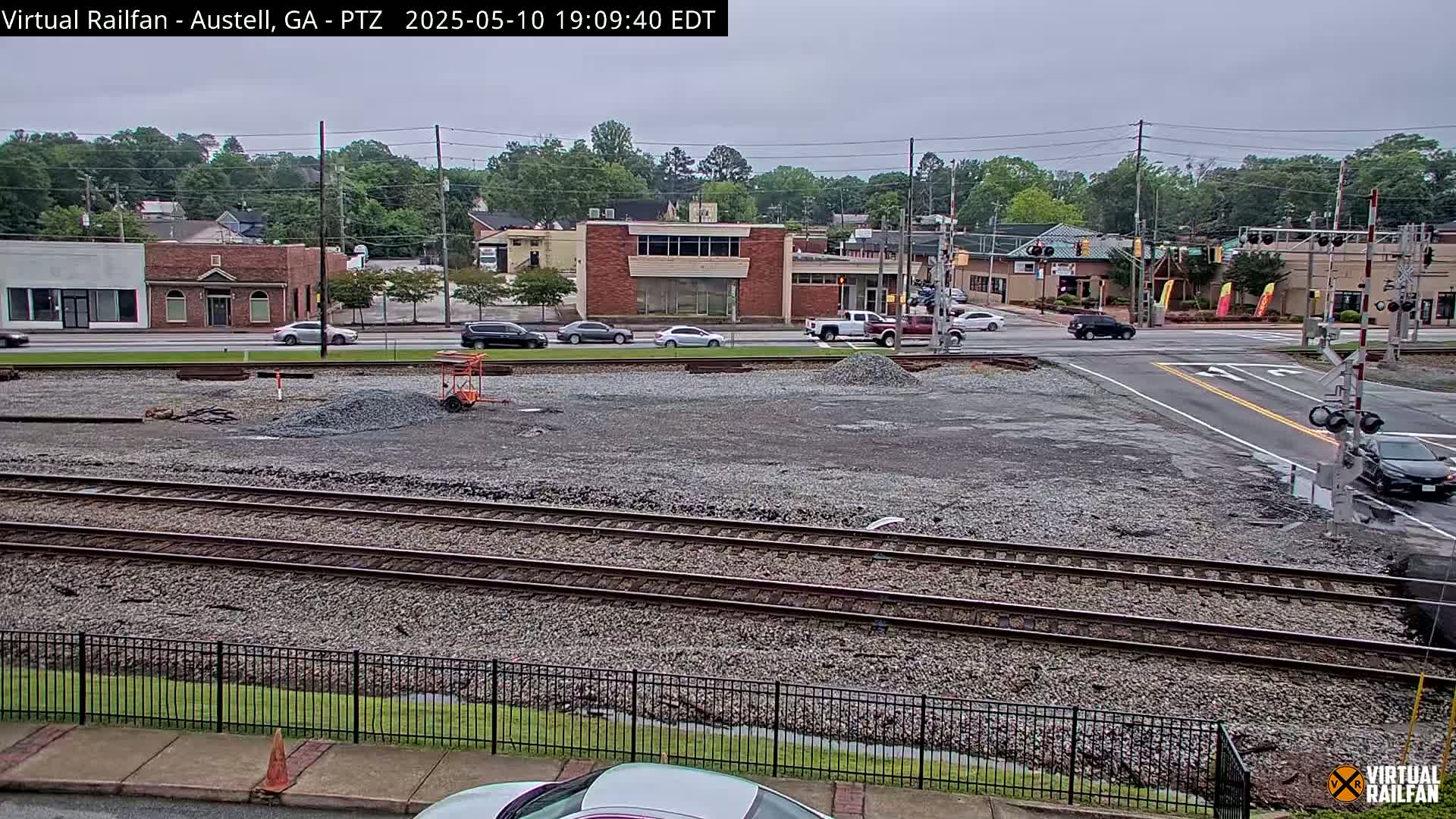 A gravel lot with railroad tracks and a metal fence sits in front of a street with several cars and buildings under an overcast sky.