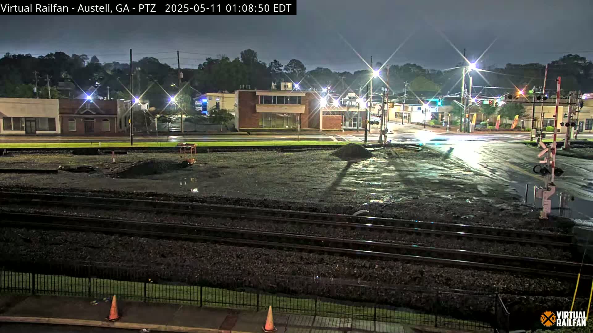 A nighttime view of a rain-slicked intersection and railroad tracks with several buildings in the background.