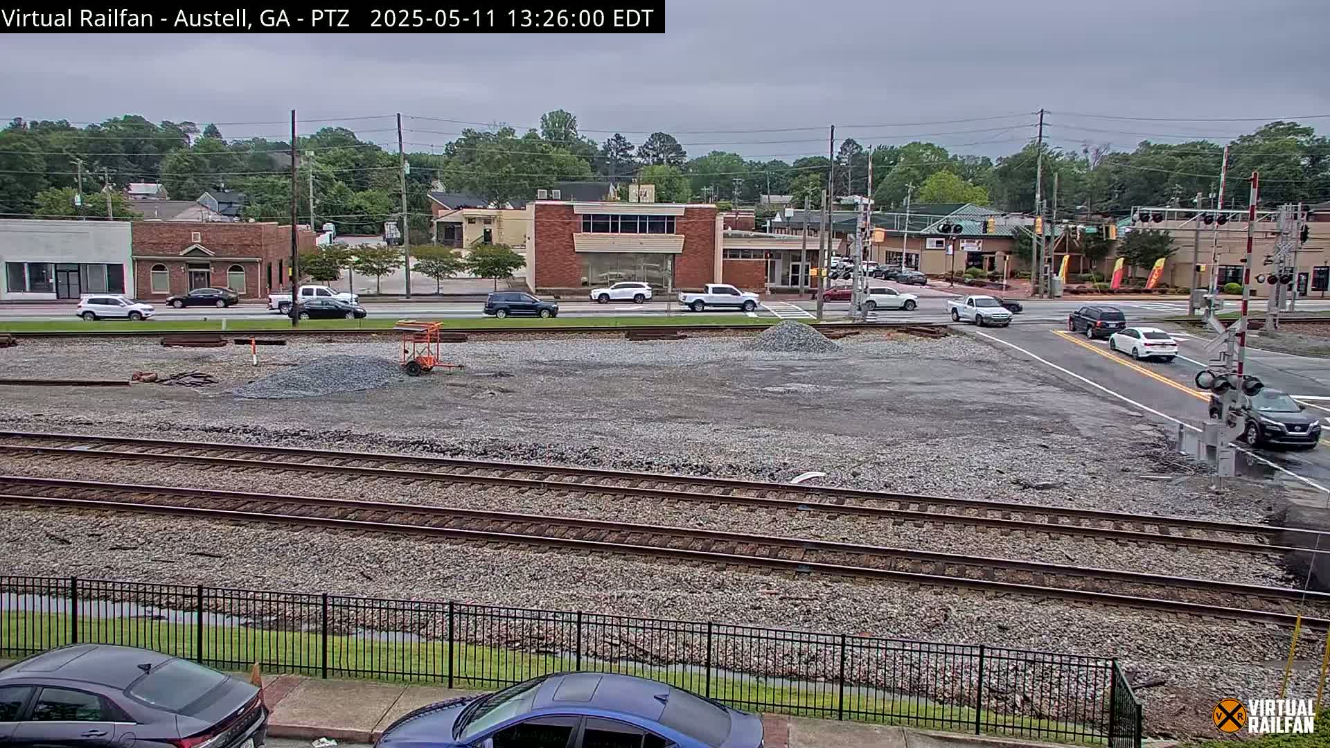 A view of a railroad crossing in a town under an overcast sky, with several vehicles at an intersection and a construction area with piles of gravel nearby.