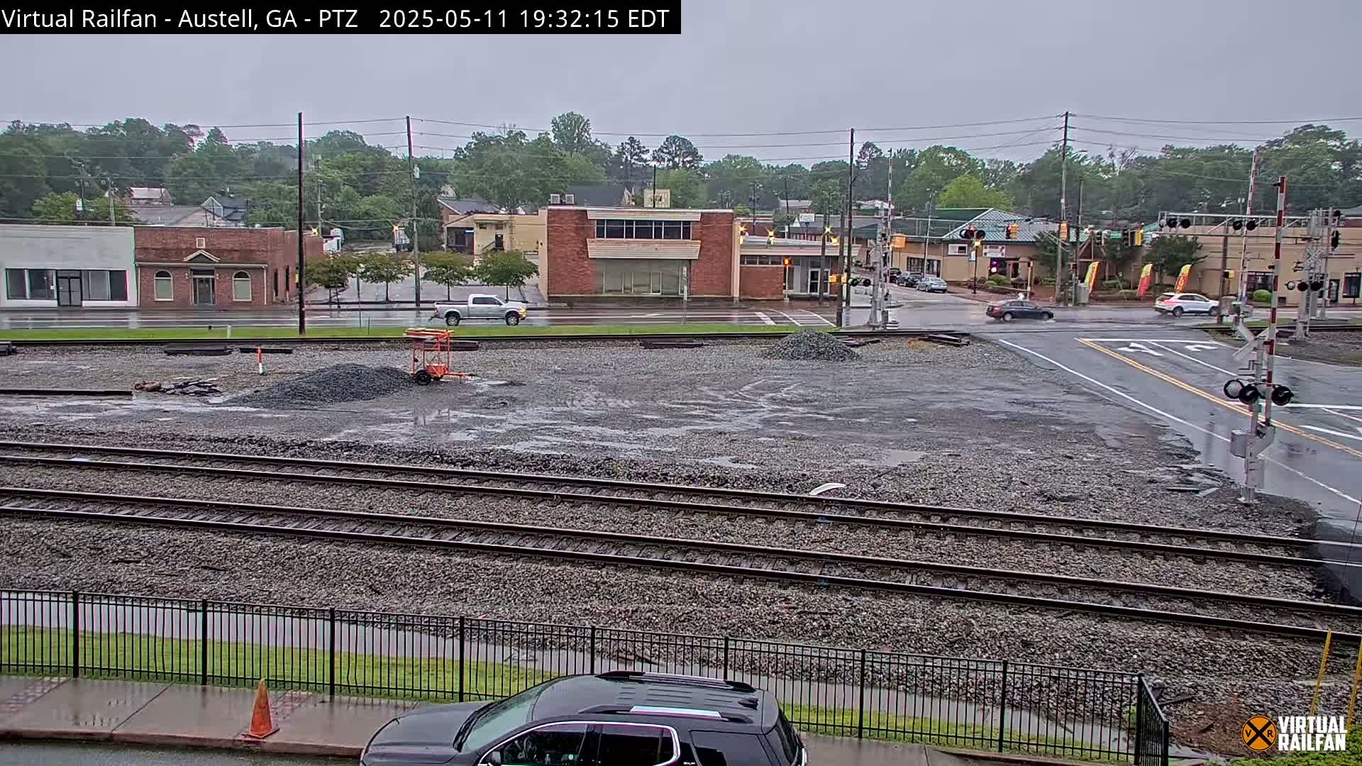 A wet, overcast day reveals a railroad crossing in a small town, with piles of gravel and construction materials near a street intersection, and several buildings in the background.