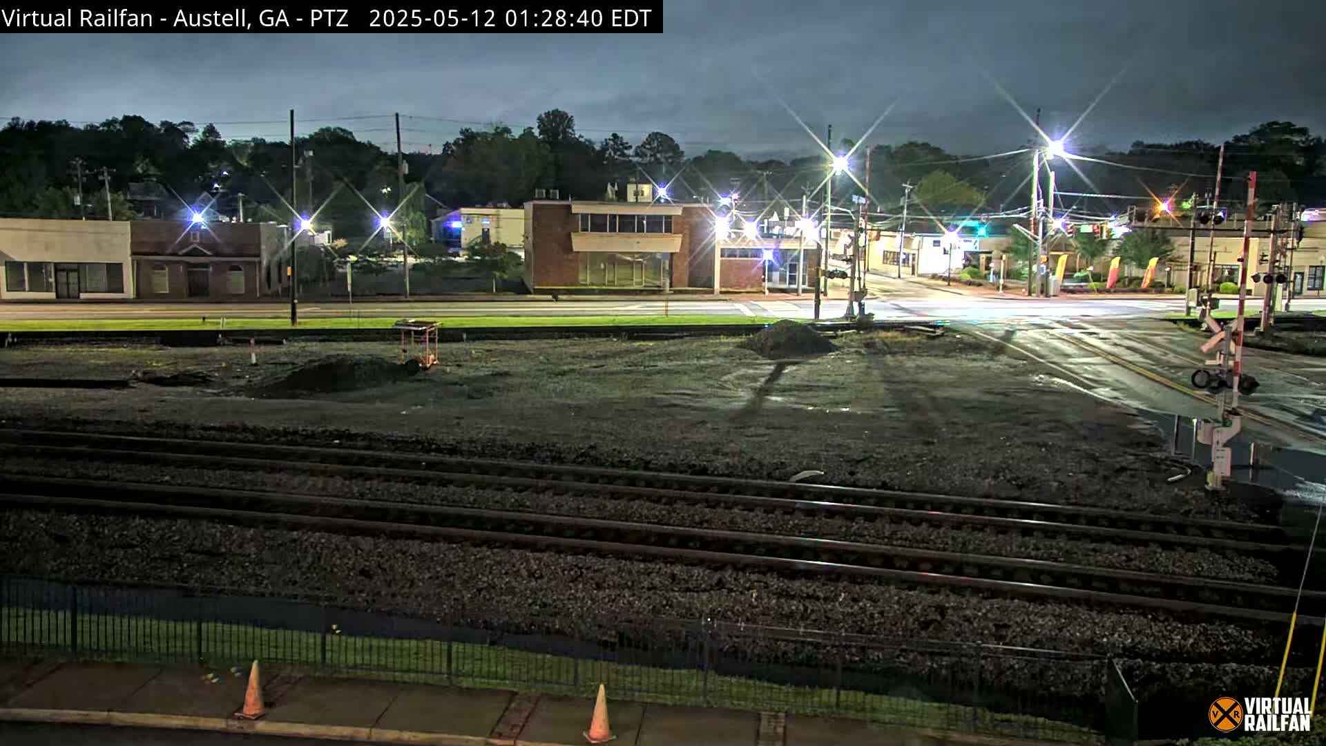 A nighttime view of a railroad crossing near a street intersection with several buildings visible in the background under a dark sky.