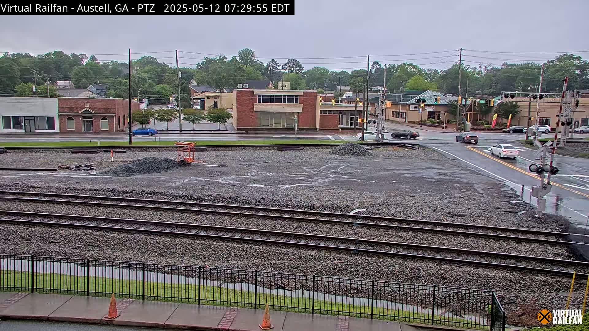 A view of railroad tracks in a town on an overcast day, with a construction site in the foreground and buildings and cars visible in the background.