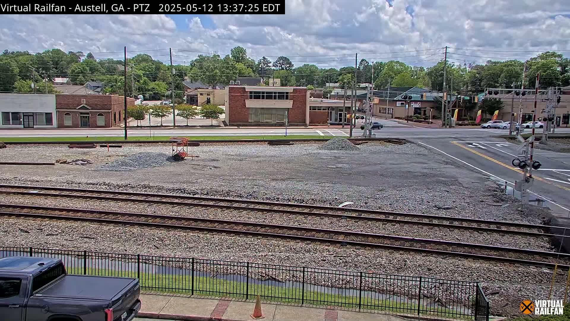 A partly cloudy day shows a view of train tracks and a gravel lot next to a street with buildings and cars, under a partly cloudy sky.