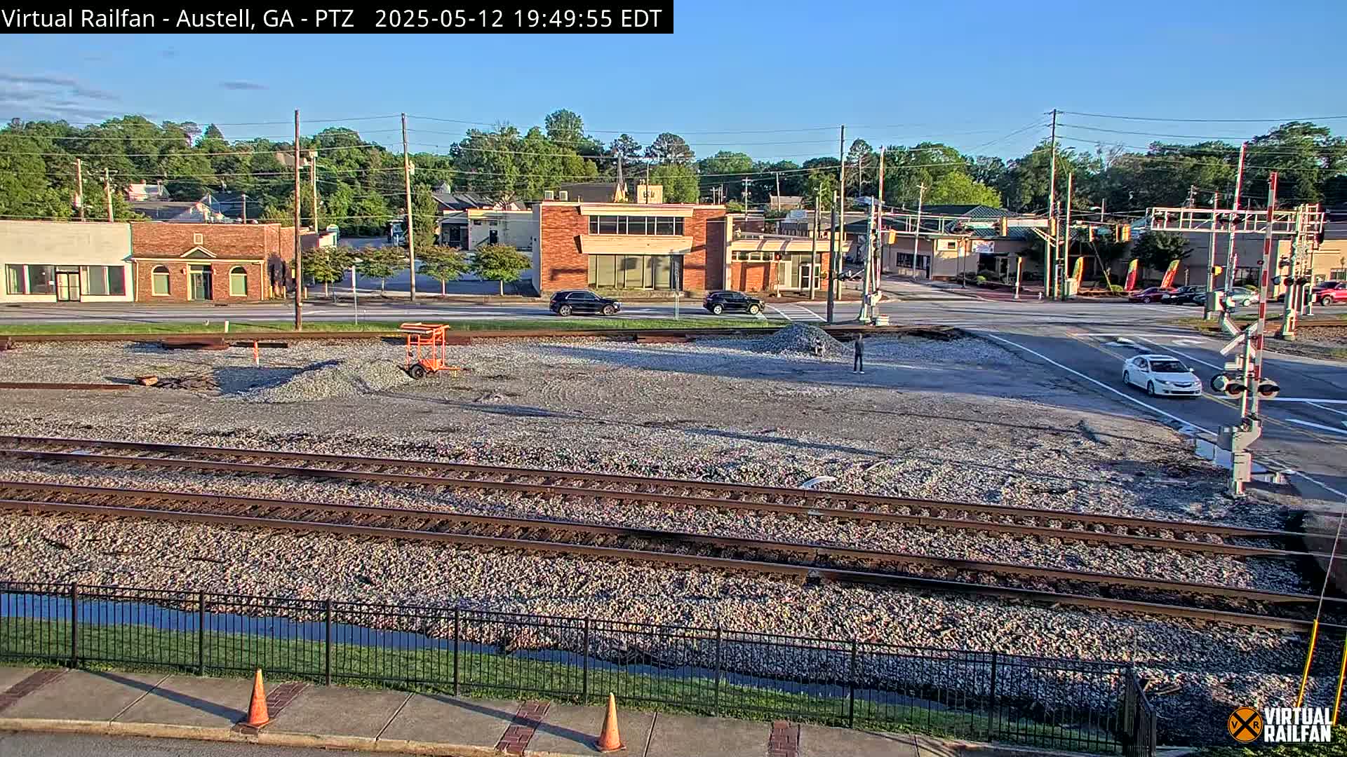 A mostly clear, daytime view shows train tracks in a gravel bed next to a road with several buildings and cars visible in the background.