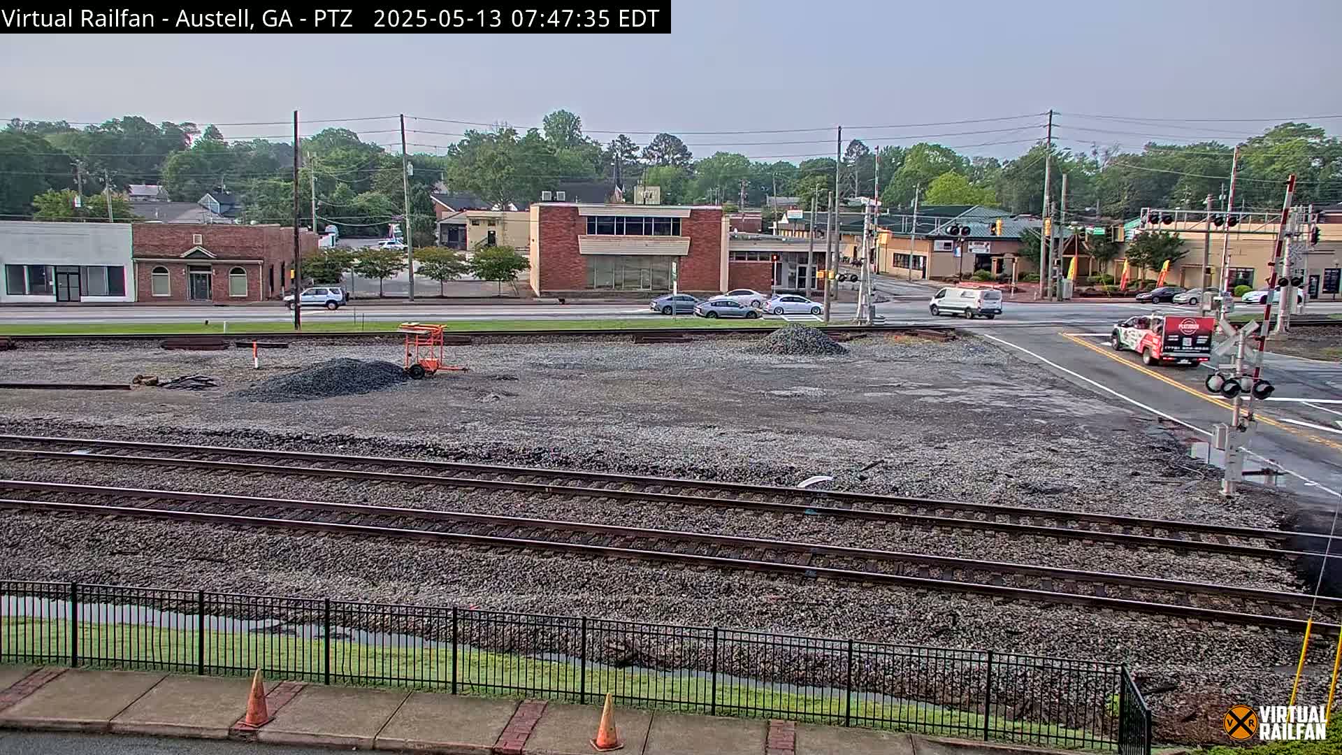 A mostly clear day reveals a railway crossing in a town, with several cars at the intersection and some construction materials on the railway tracks.