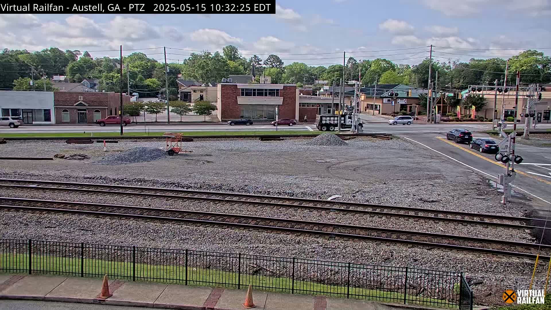 A partly cloudy day reveals a construction site next to railroad tracks and a street intersection, with several vehicles visible on the road and a few buildings in the background.