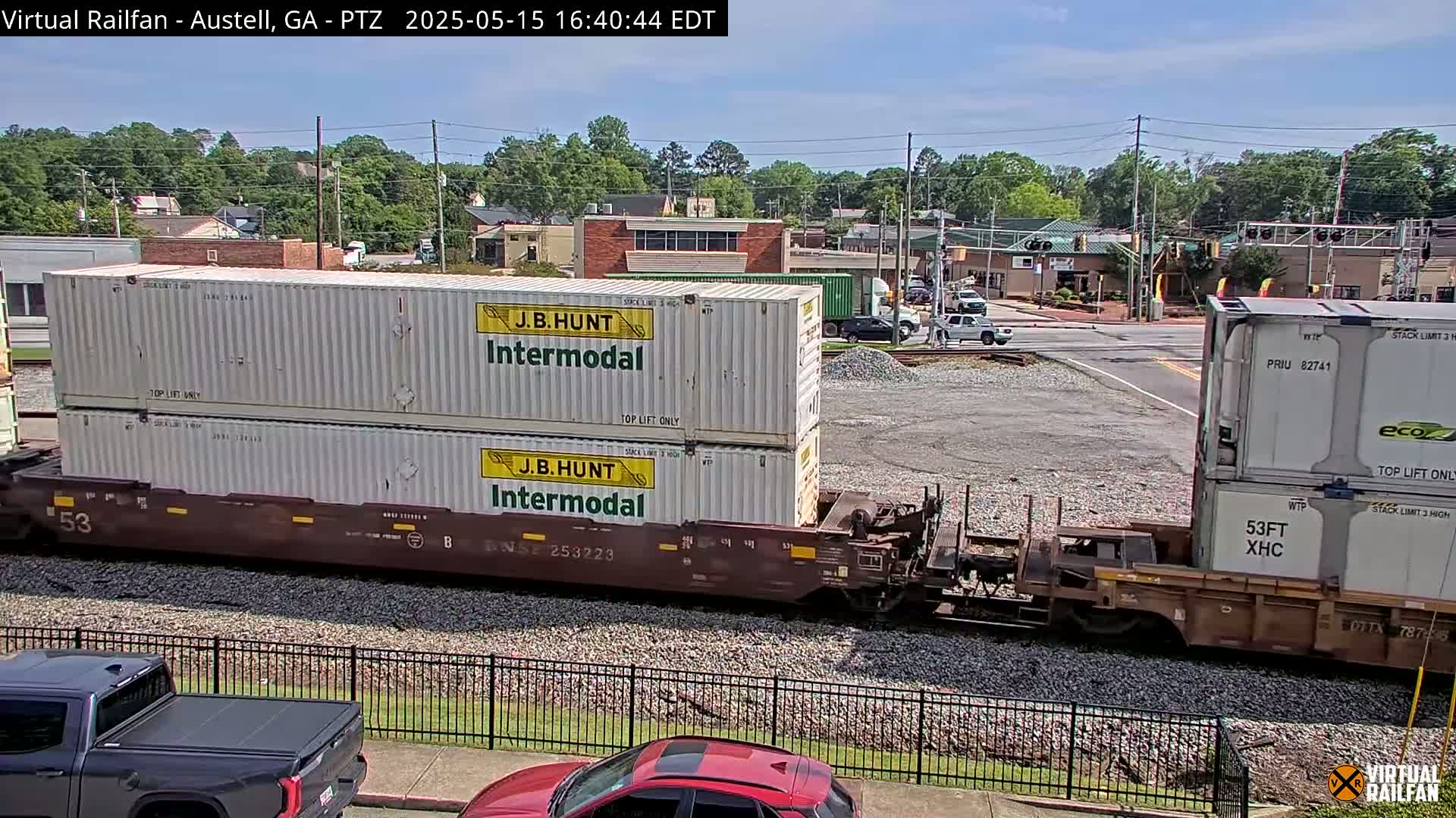 A train carrying two stacked shipping containers labeled "J.B. Hunt Intermodal" is seen passing through a town under a mostly sunny sky.