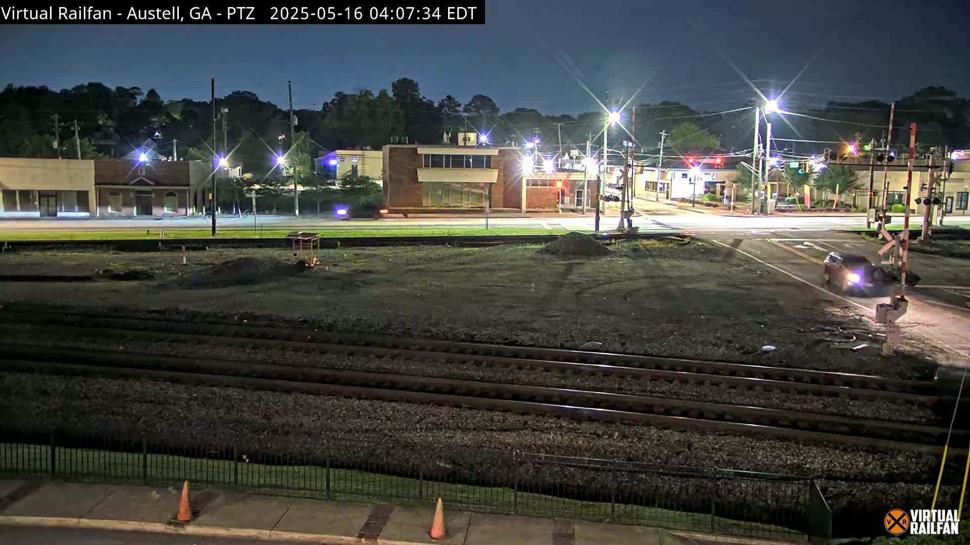 A nighttime view of a street intersection with railroad tracks, buildings, and a single vehicle at the crossing.