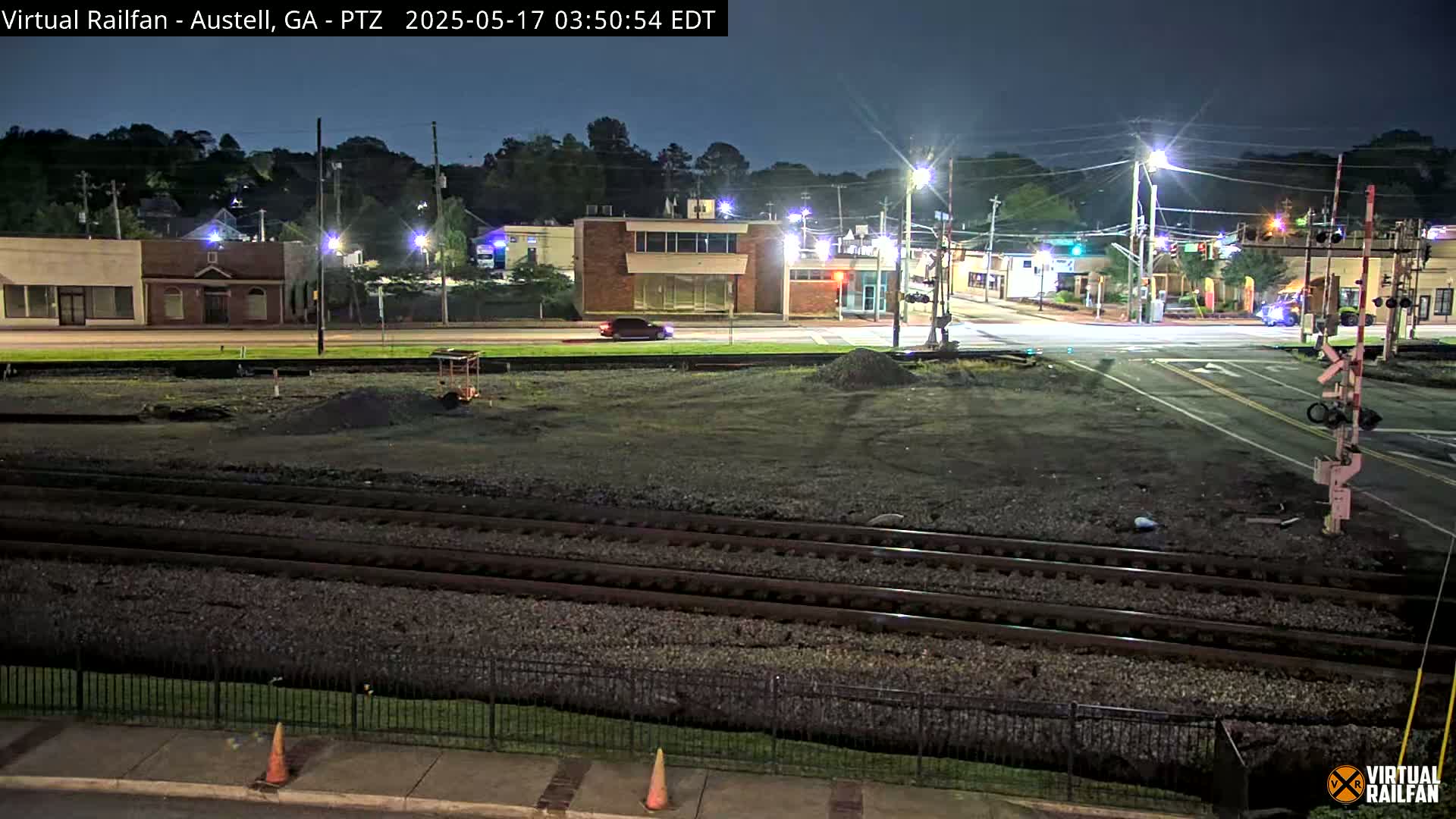 A nighttime view of a street intersection with railroad tracks in the foreground, featuring several buildings under streetlights.
