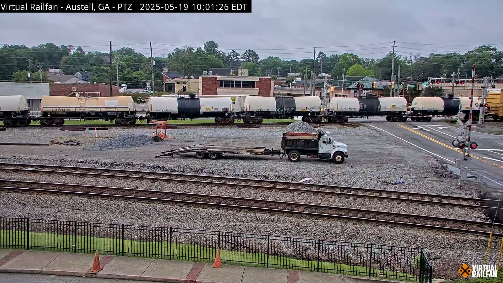 A line of tank cars sits on a railway crossing next to a construction site with a dump truck and a flatbed trailer under an overcast sky.