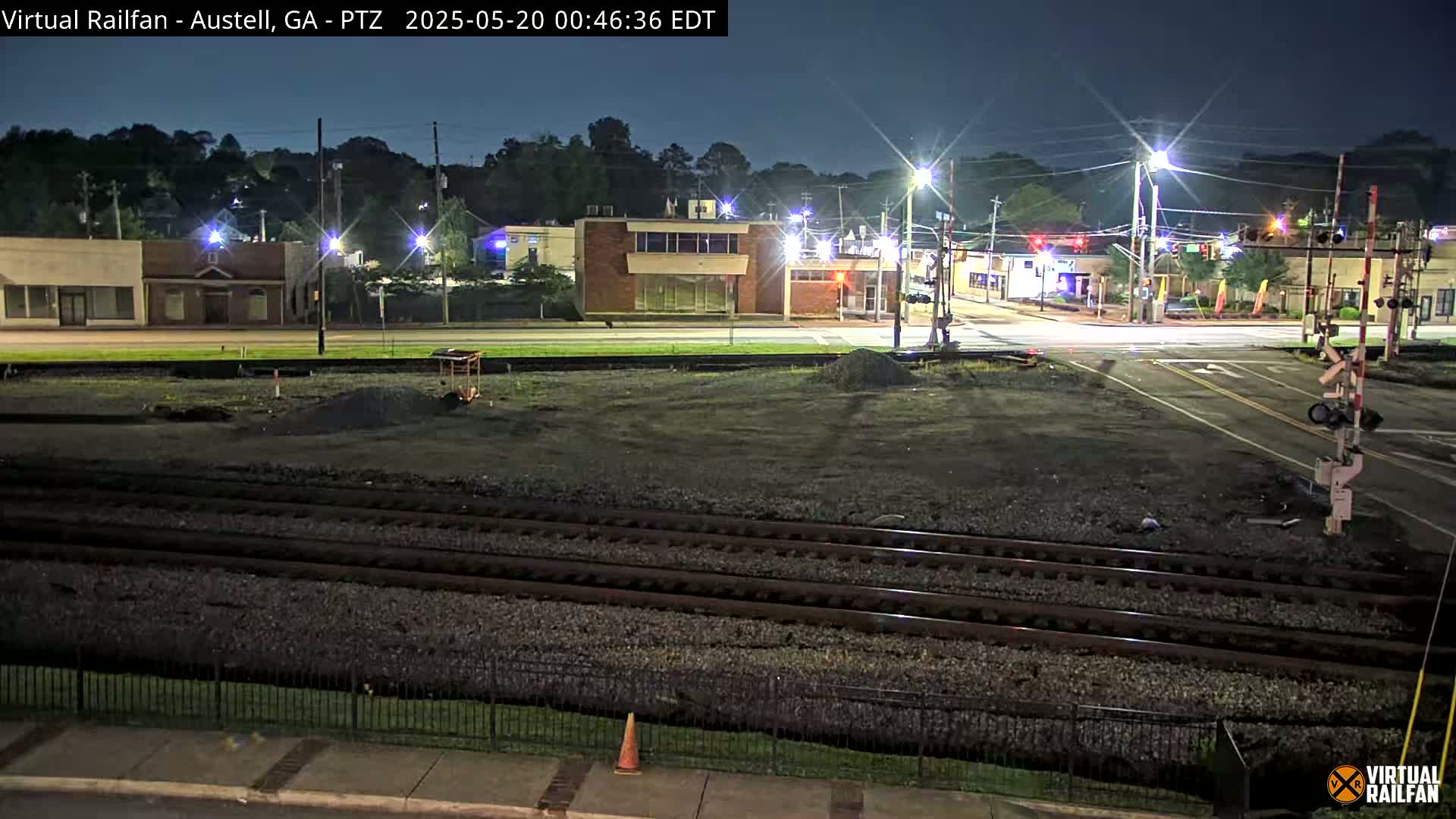 A nighttime view of a railroad crossing near a street intersection with several buildings visible in the background under a clear sky.
