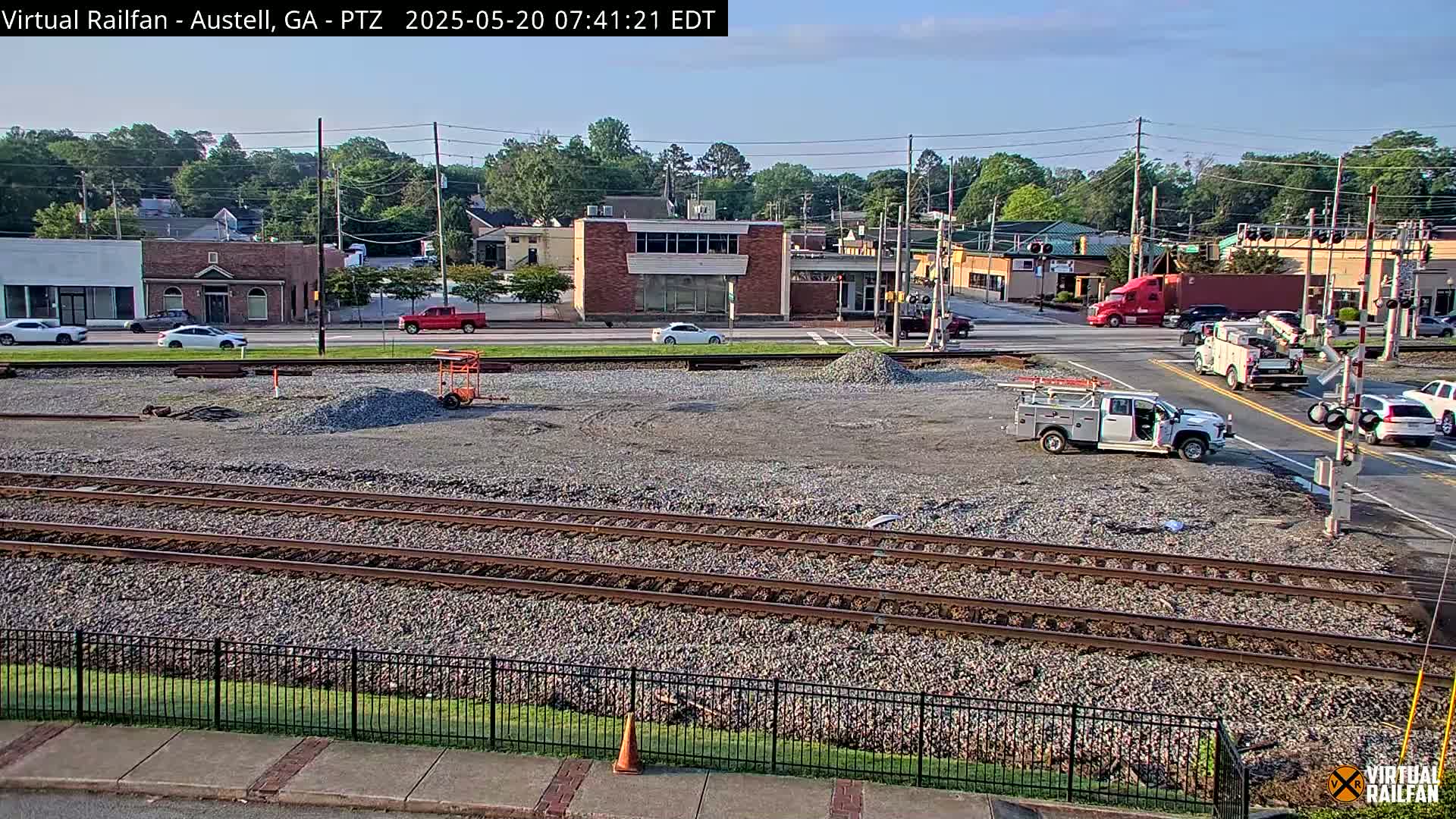 A view of a mostly empty gravel lot next to train tracks, with several vehicles and buildings visible in the background under a partly sunny sky.