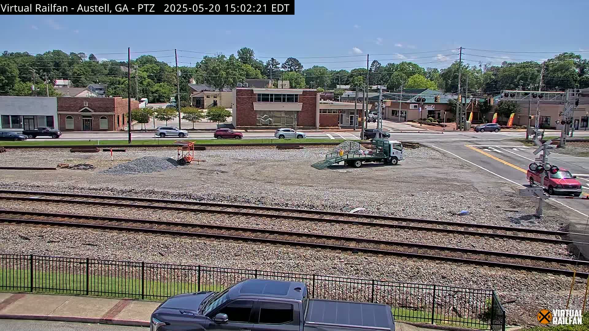 Under a sunny sky, a view of railroad tracks next to a town's street intersection shows several vehicles, construction equipment, and buildings.