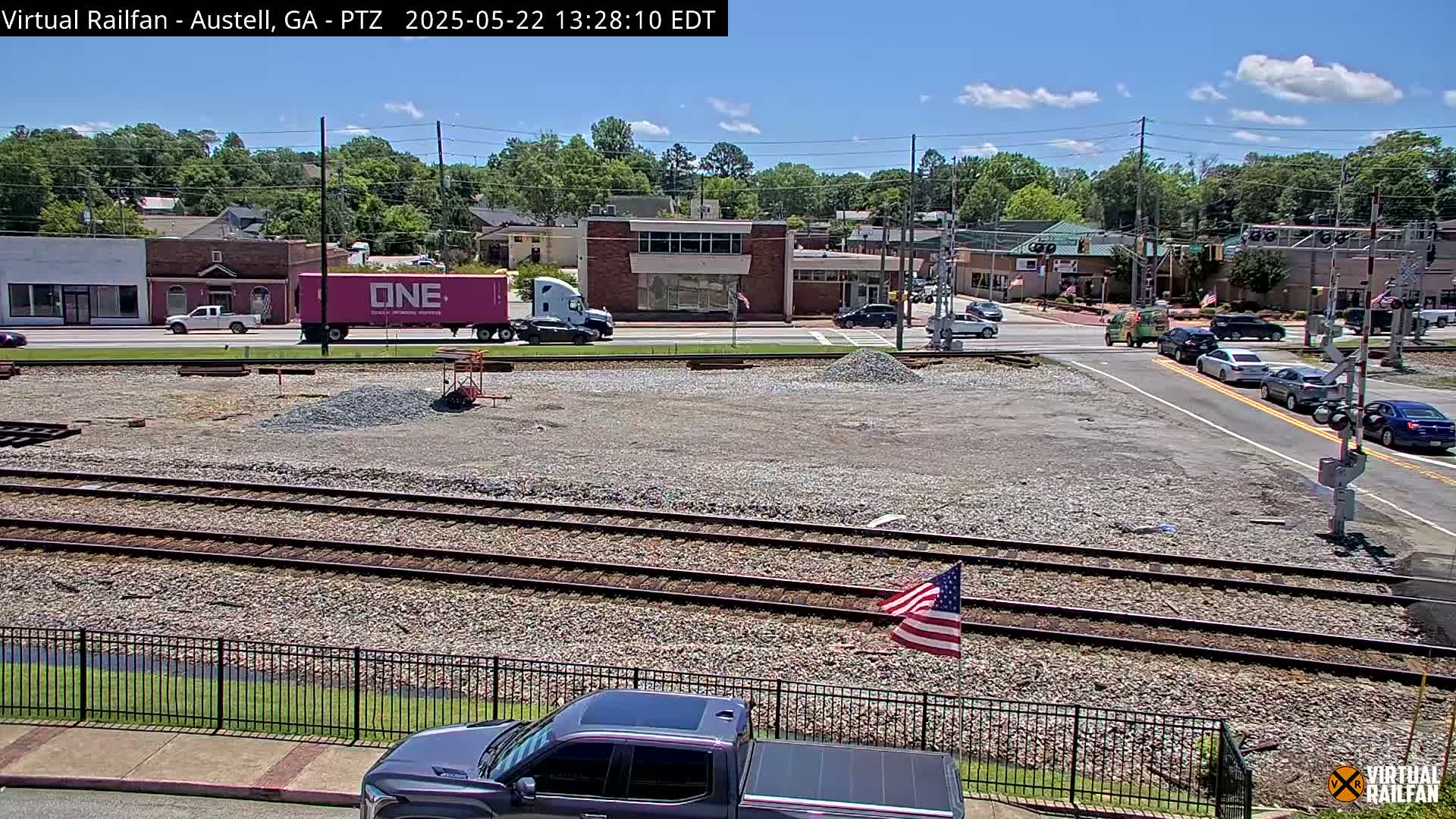 A sunny day reveals a view of train tracks bordered by a fence, a gravel area with construction materials, and several vehicles at a nearby intersection, with buildings and trees in the background.
