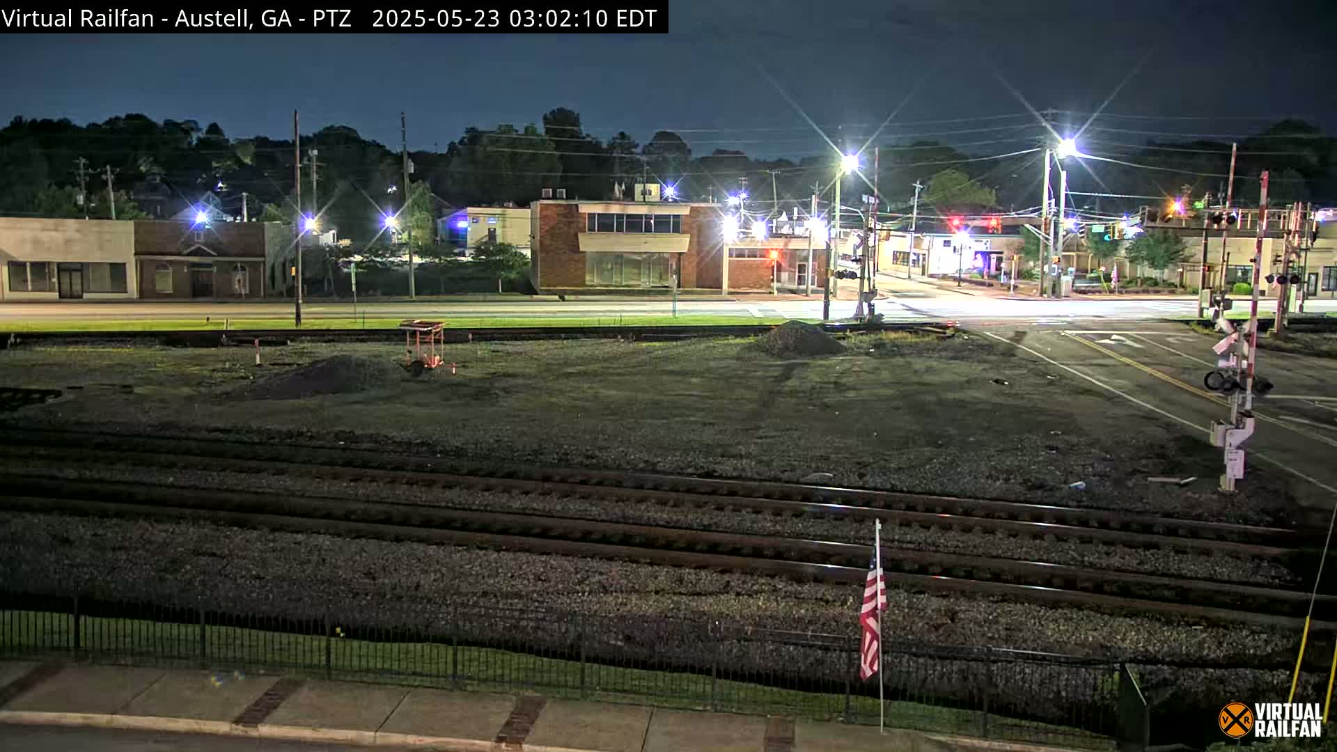 A nighttime view of a railroad crossing near a street intersection with several buildings visible under a clear sky.
