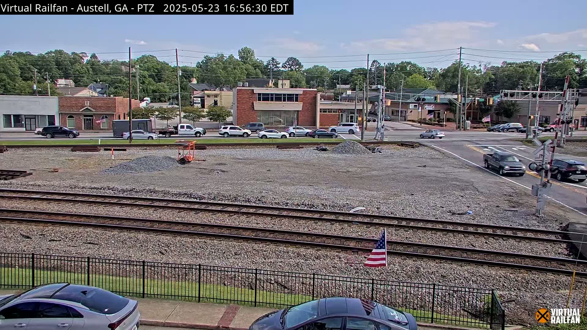 A mostly clear day view of a train track crossing in a small town, with several cars parked and driving near a construction site.