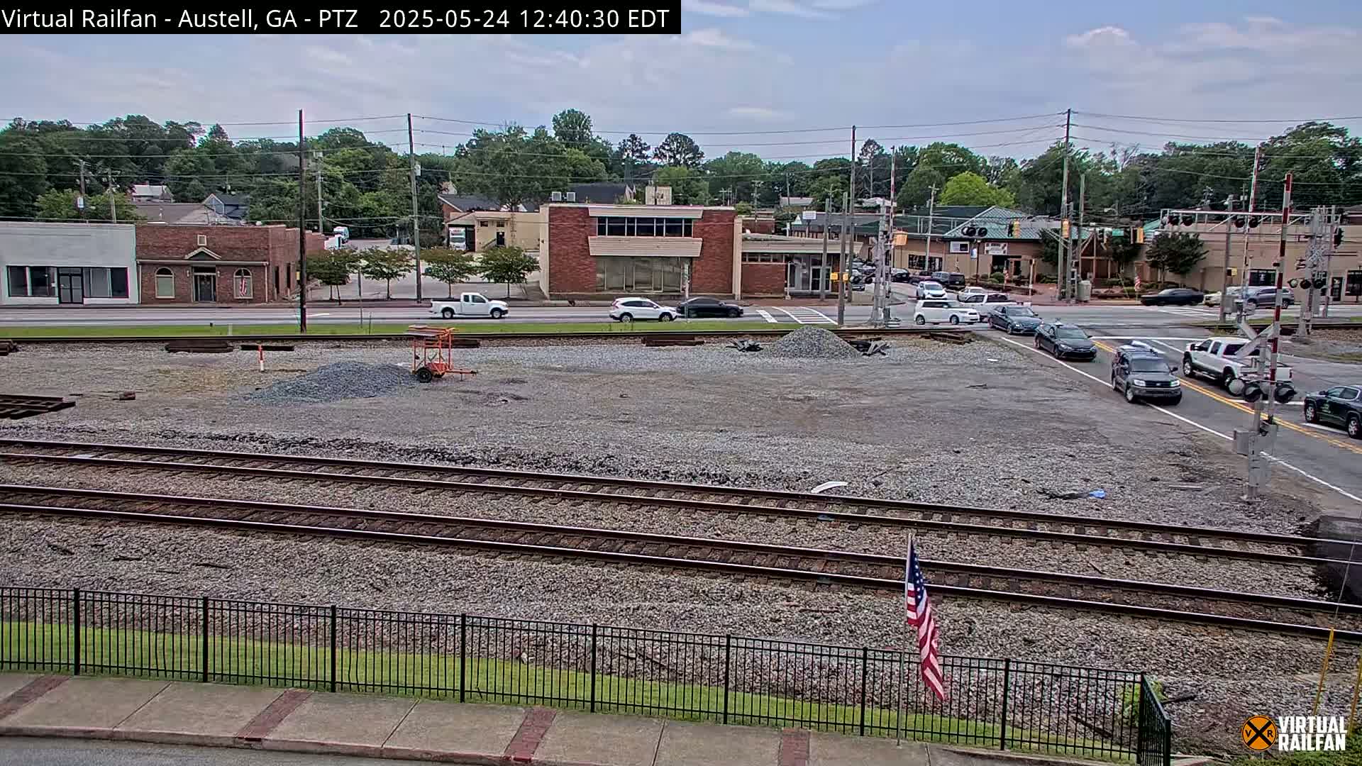 A mostly sunny day reveals a train track bed with gravel, bordered by a fence, in front of a street intersection with several vehicles and buildings.