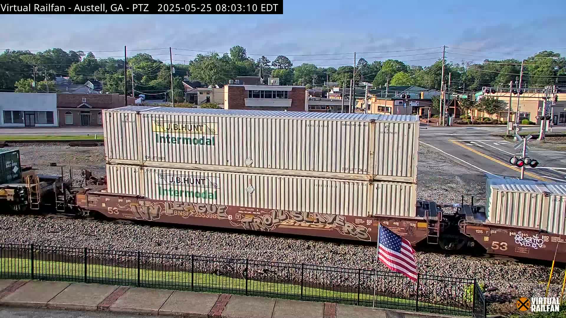 A freight train carrying stacked shipping containers sits on a railway track near a road intersection in a town, under a mostly clear sky.
