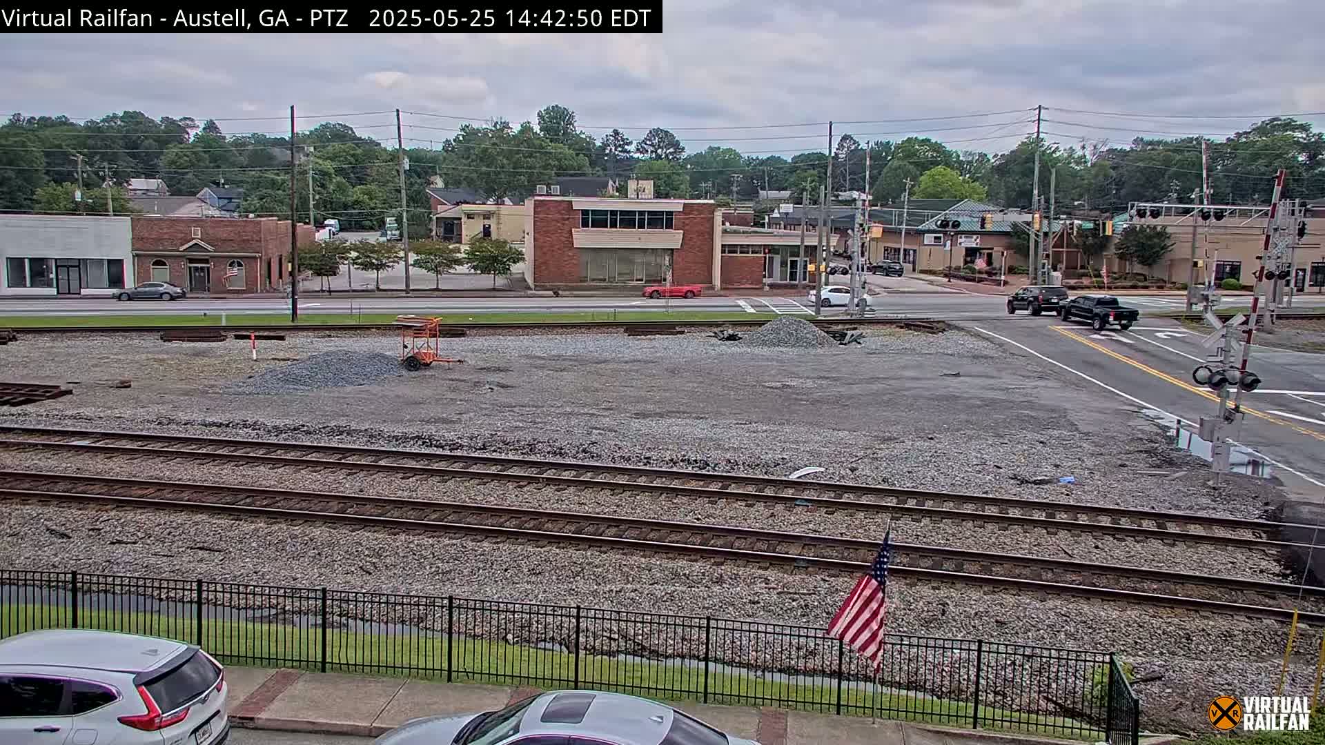 A mostly cloudy day shows a view of railroad tracks next to a street intersection in a small town, with a few cars and trucks visible, and some construction materials piled on the tracks.